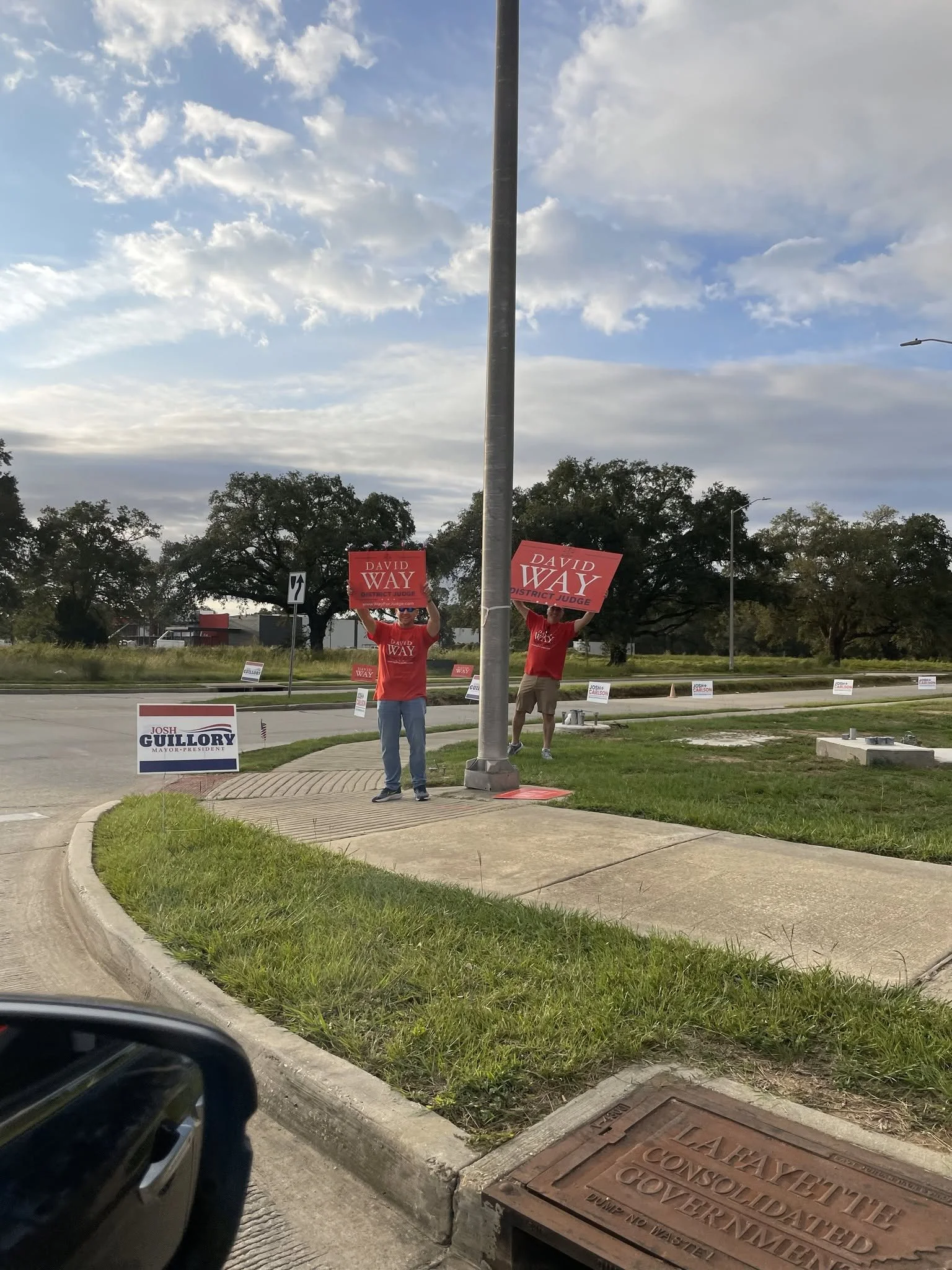 People holding campaign signs that say 'David Way District Judge' standing near a light pole and road with signs supporting Josh Guillory for mayor, a manhole cover reading 'GOVERNMENT ALLEY' in a grassy area, and trees in the background.