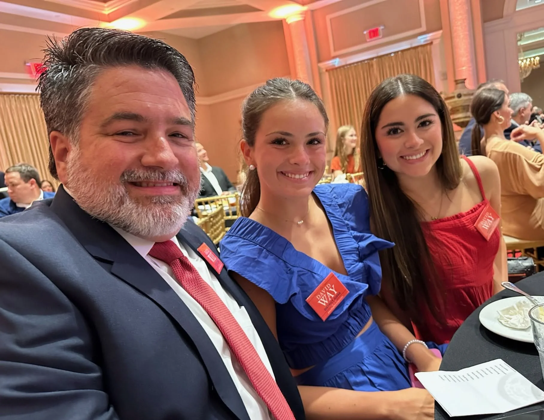 Three people seated at a formal event, smiling at the camera. The man on the left is wearing a dark suit, white shirt, red tie, and has a gray beard. The woman in the middle has brown hair tied back, wearing a blue dress with ruffled sleeves. The wom