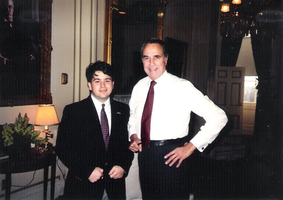 Two men in formal attire standing indoors, one younger with dark hair and the other older with gray hair, in a decorated room with curtains, a lamp, and framed artwork.