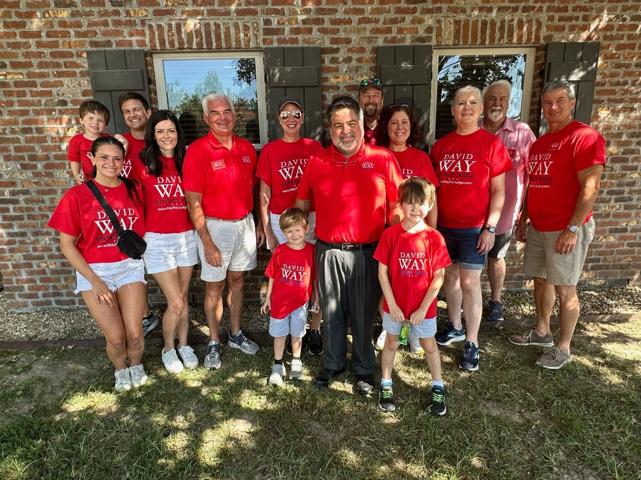 A group of people, including children and adults, posing outdoors in front of a brick building, all wearing red campaign shirts promoting David Way for district judge.
