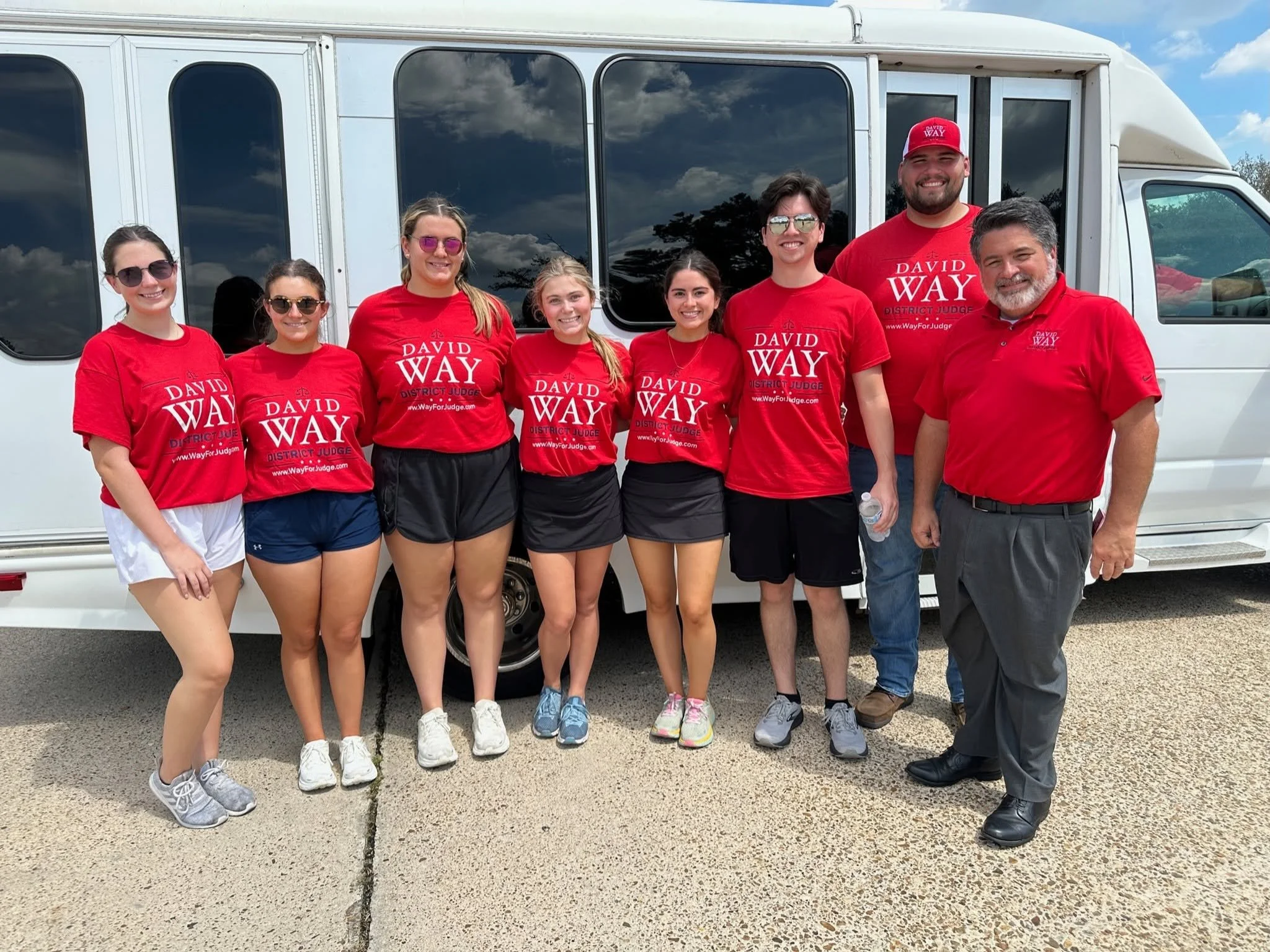 Group of eight people standing in front of a white bus, all wearing red shirts with campaign slogans, smiling, with cloudy sky reflecting on bus windows.