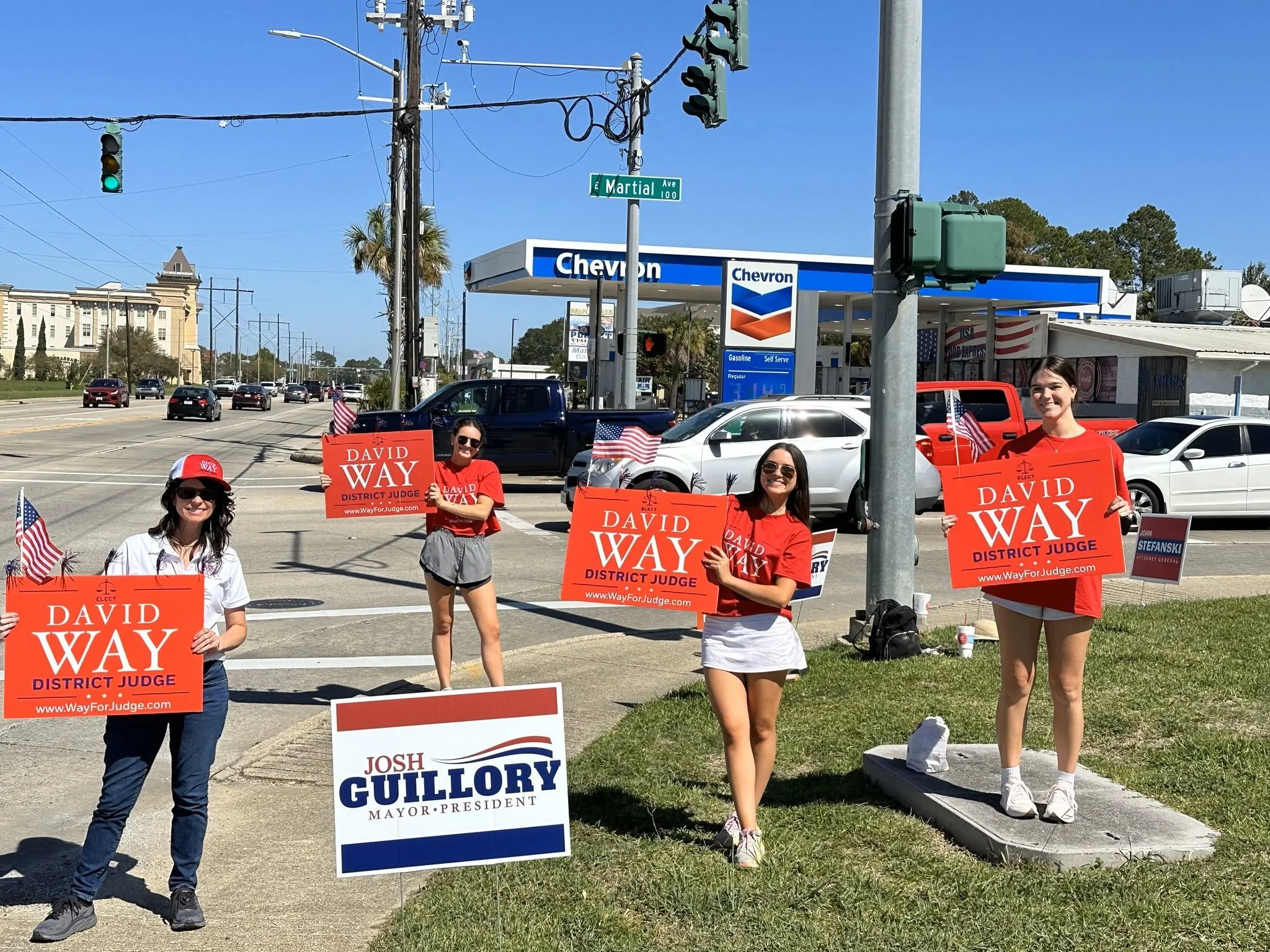 A group of people supporting political candidates, holding campaign signs, and standing near a Chevron gas station on a sunny day with clear blue skies. The signs promote candidates David Way, Josh Guillory, and others for district judge and mayor or