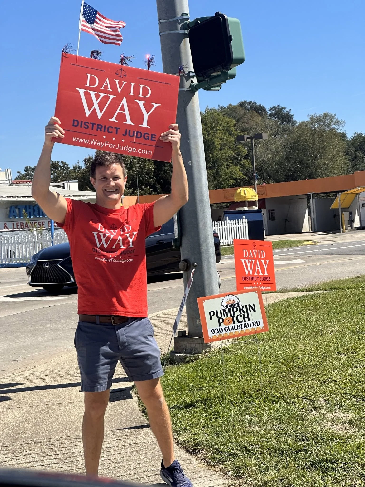 A man in a red T-shirt and shorts holding a campaign sign that reads 'David Way District Judge' and a website URL, standing on a sidewalk near campaign signs and a pole with an American flag and other signs, during daytime with clear blue skies.