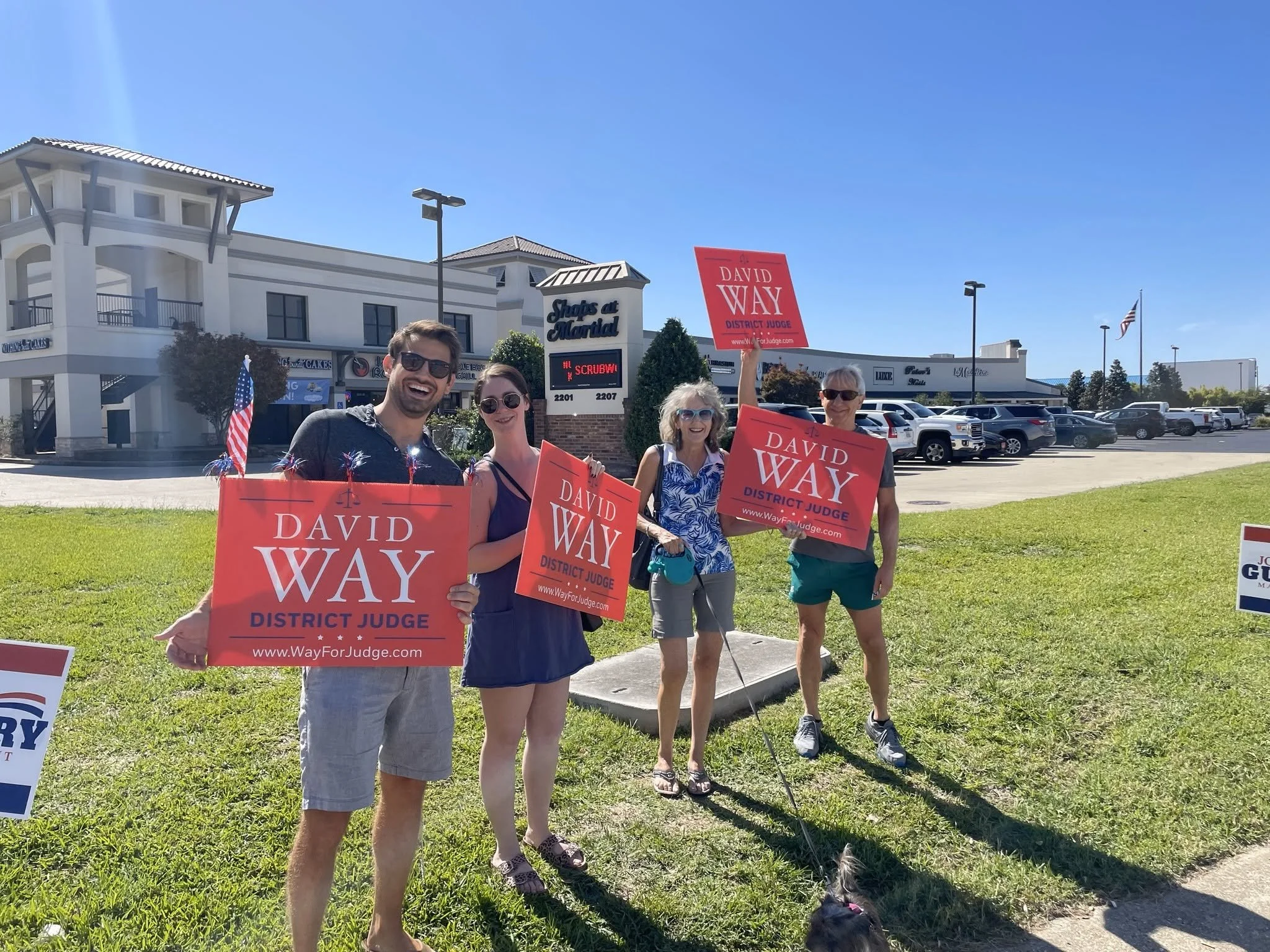 Group of people holding campaign signs for David Way, District Judge, standing in a parking lot in front of a shopping center on a sunny day.