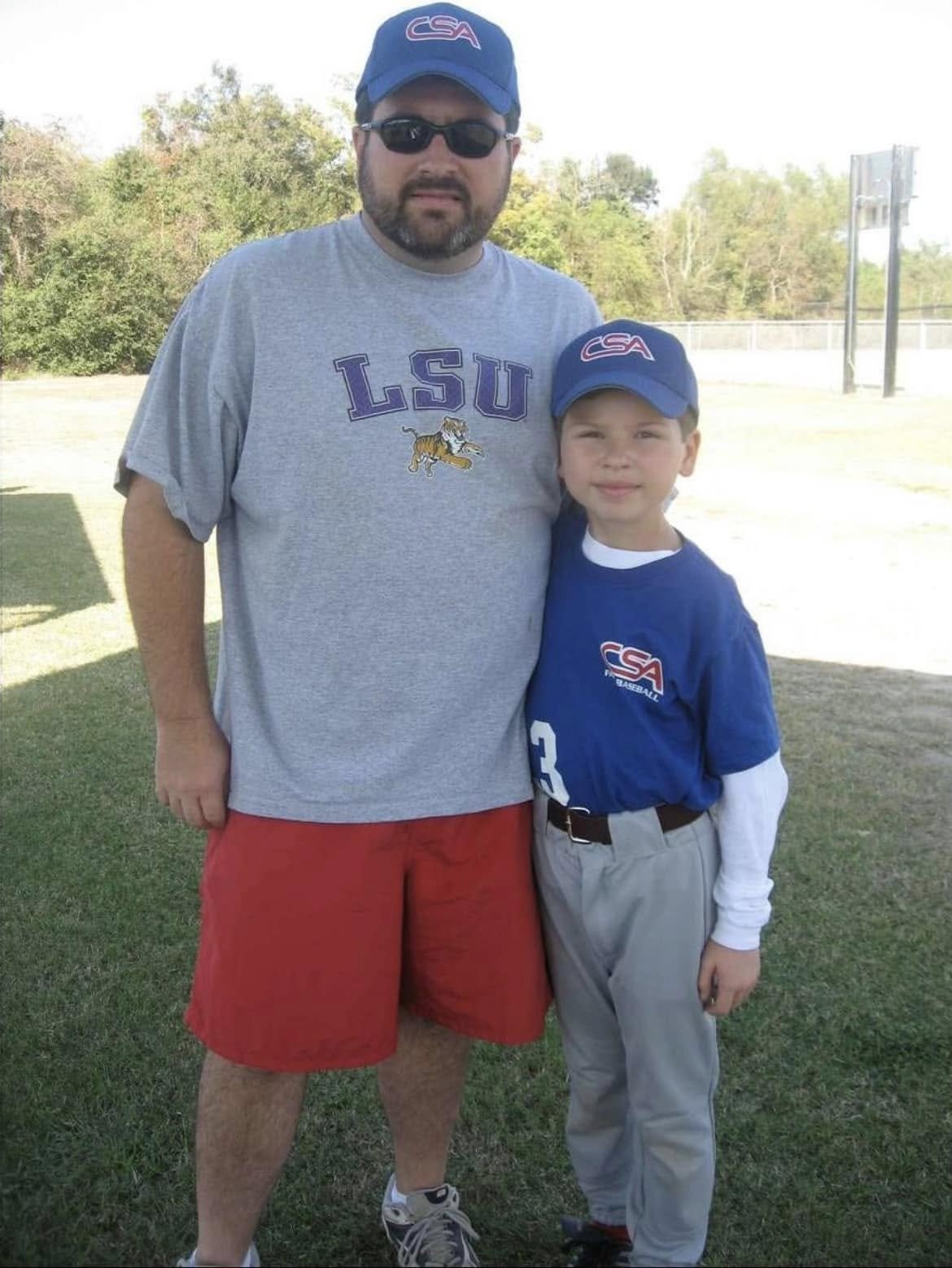 A man and a boy standing outdoors, both wearing baseball caps with the USA logo and team jerseys, posing for a photo on a grassy field with trees and sports field equipment in the background.