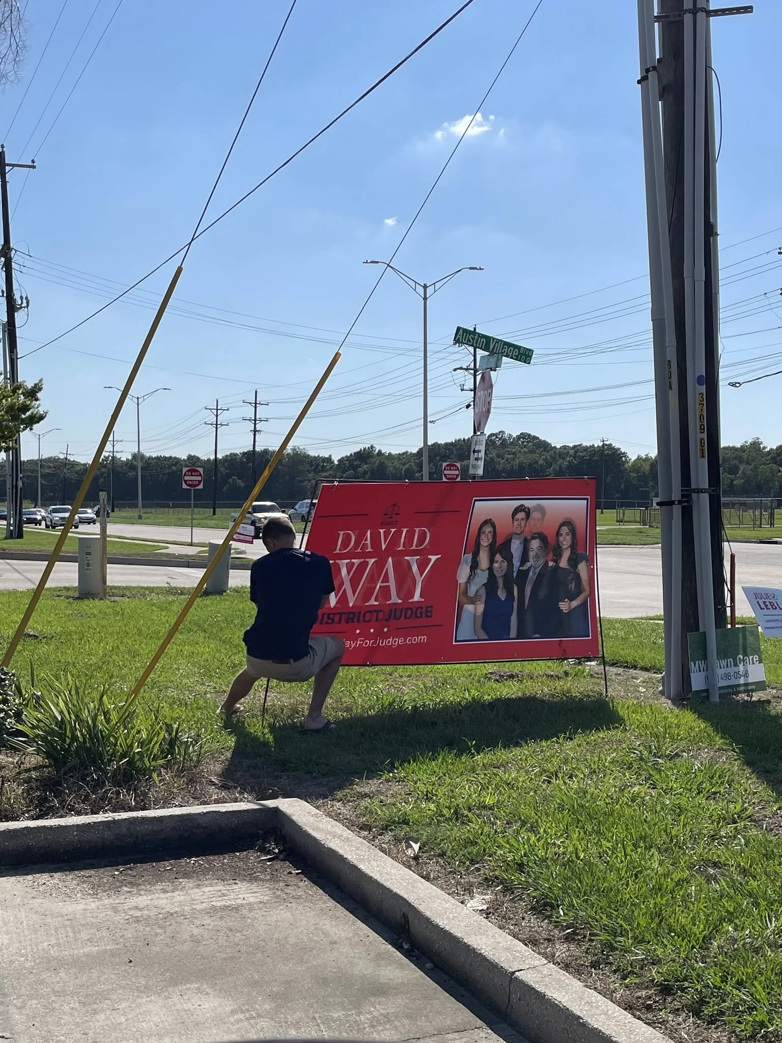 A political campaign sign for David Wayne, District Judge, with a group of six young people pictured on the sign. The scene is outdoors on a bright, clear day with power lines, street signs, and parked cars in the background. A man is kneeling in fro