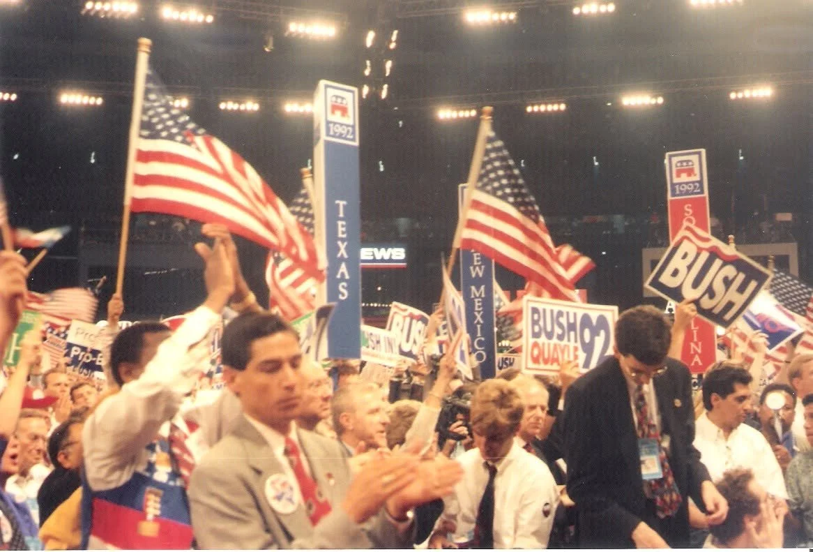 A crowd at a political rally, waving American flags and holding signs supporting George W. Bush for president in 2000 and 2004, inside a large indoor arena.