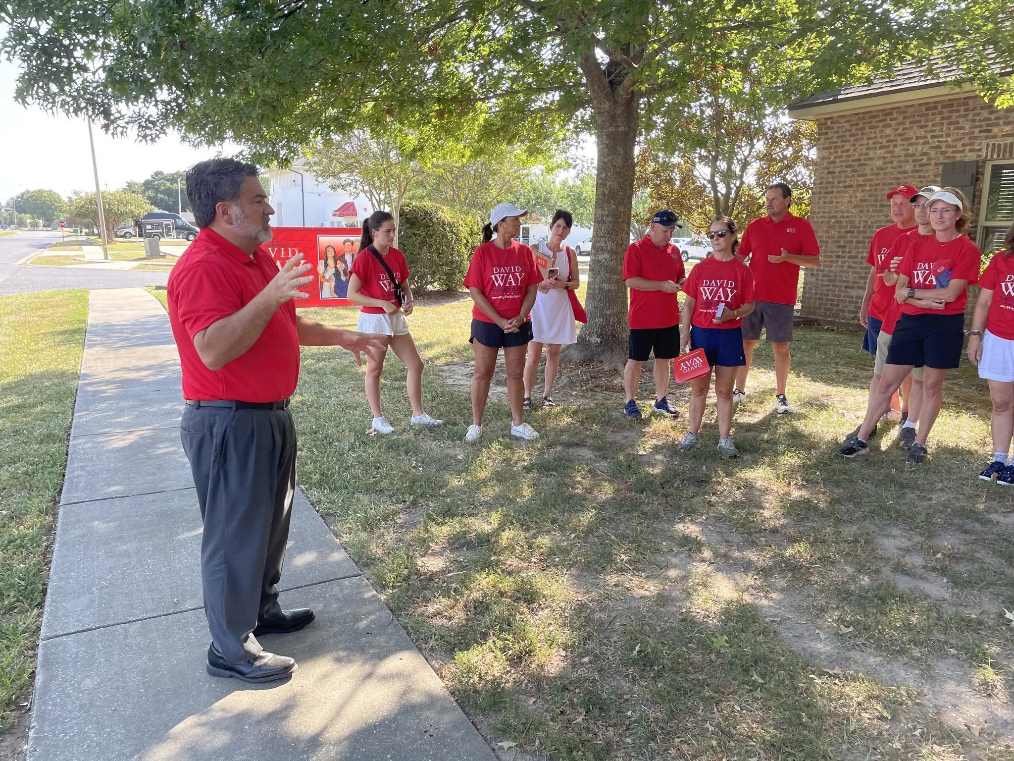 A group of people wearing red t-shirts gathered outdoors under a large tree, listening to a man speaking. The setting appears to be near a building with brick walls and some campaign posters.