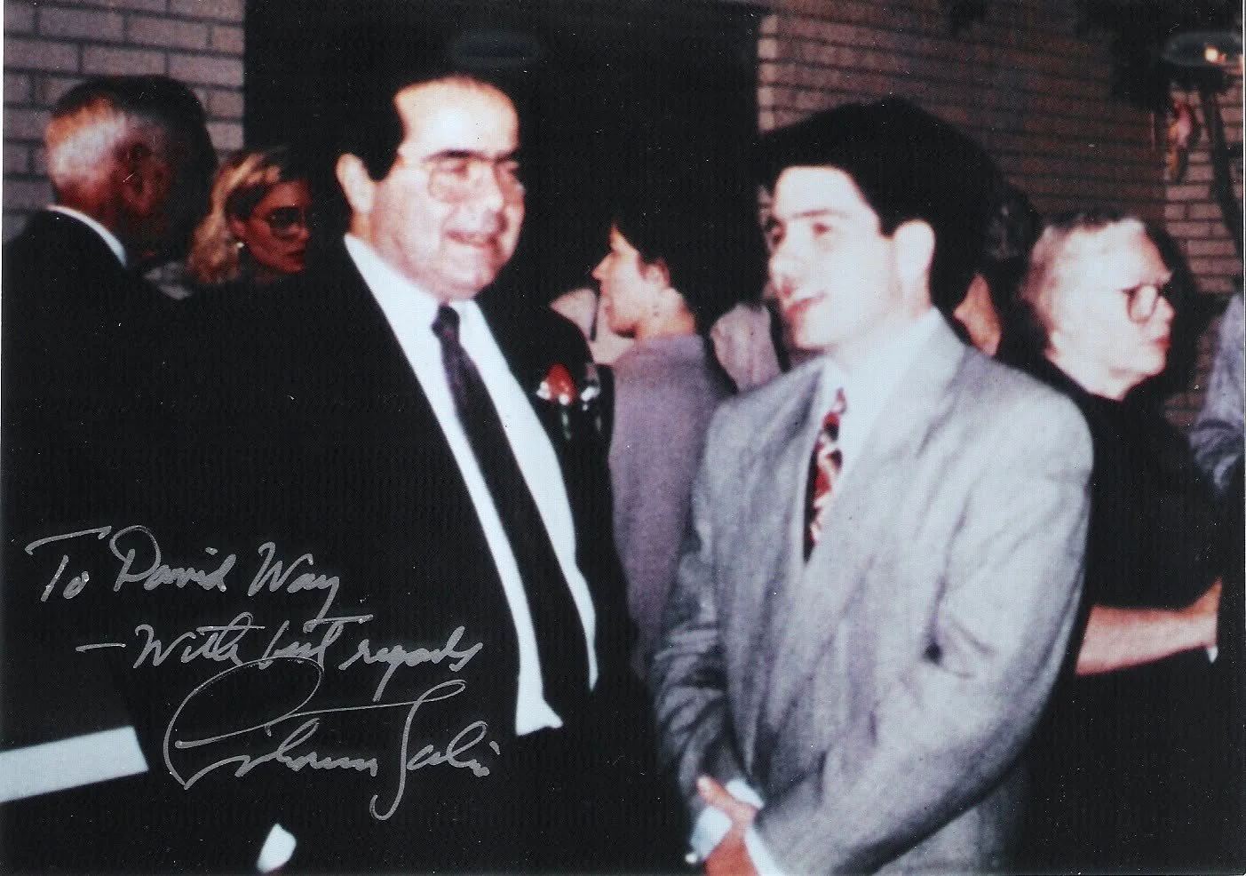 Two men in suits engaged in conversation at a social event, with women and others in the background. One of the men is older with dark hair, the other younger with dark hair and glasses. The image includes a handwritten note wishing someone a happy b