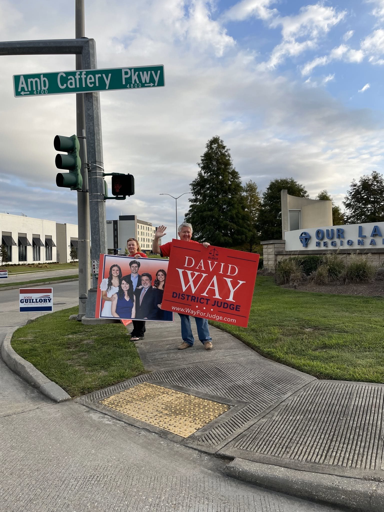 People holding campaign signs and waving on sidewalk at street corner with traffic light, street signs, and a building in the background.