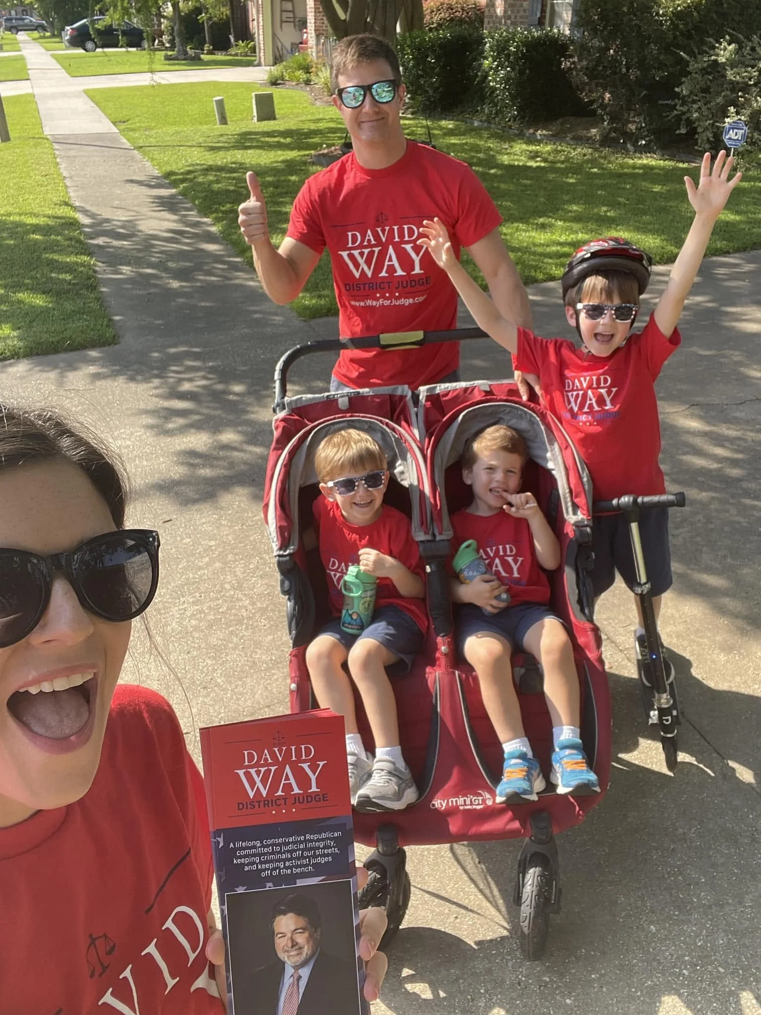 A woman taking a selfie with a man and three children outdoors on a sunny day. The man and children are wearing red campaign shirts supporting David Way, a district judge candidate. The woman is wearing sunglasses, and the children are sitting in a d