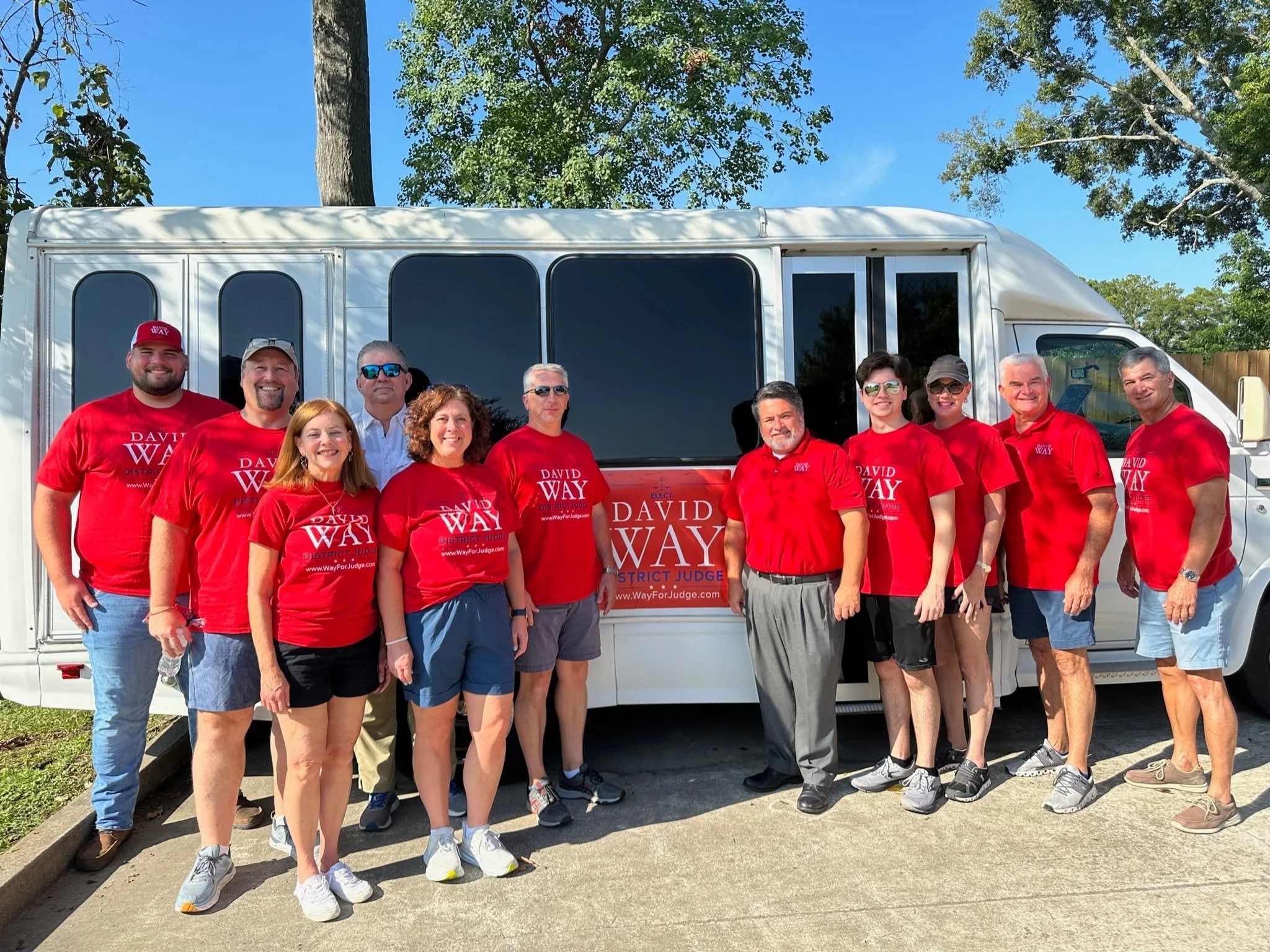 Group of people standing in front of a large vehicle, wearing red shirts with campaign slogans, smiling, with trees and blue sky in background.