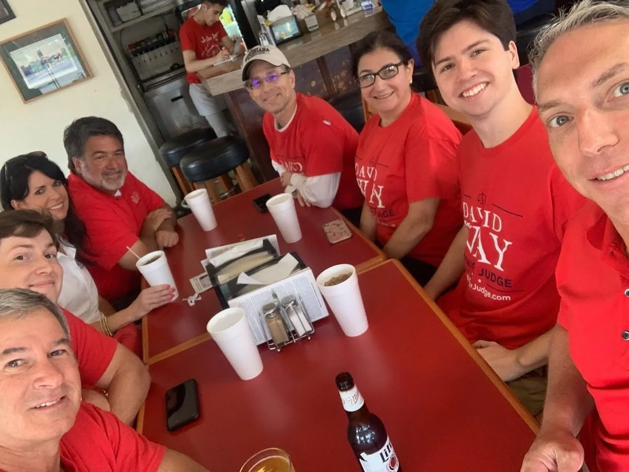 Group of people sitting around a red table in a restaurant, smiling, wearing red shirts, some with sunglasses, with drinks and condiments, and a bar in the background.