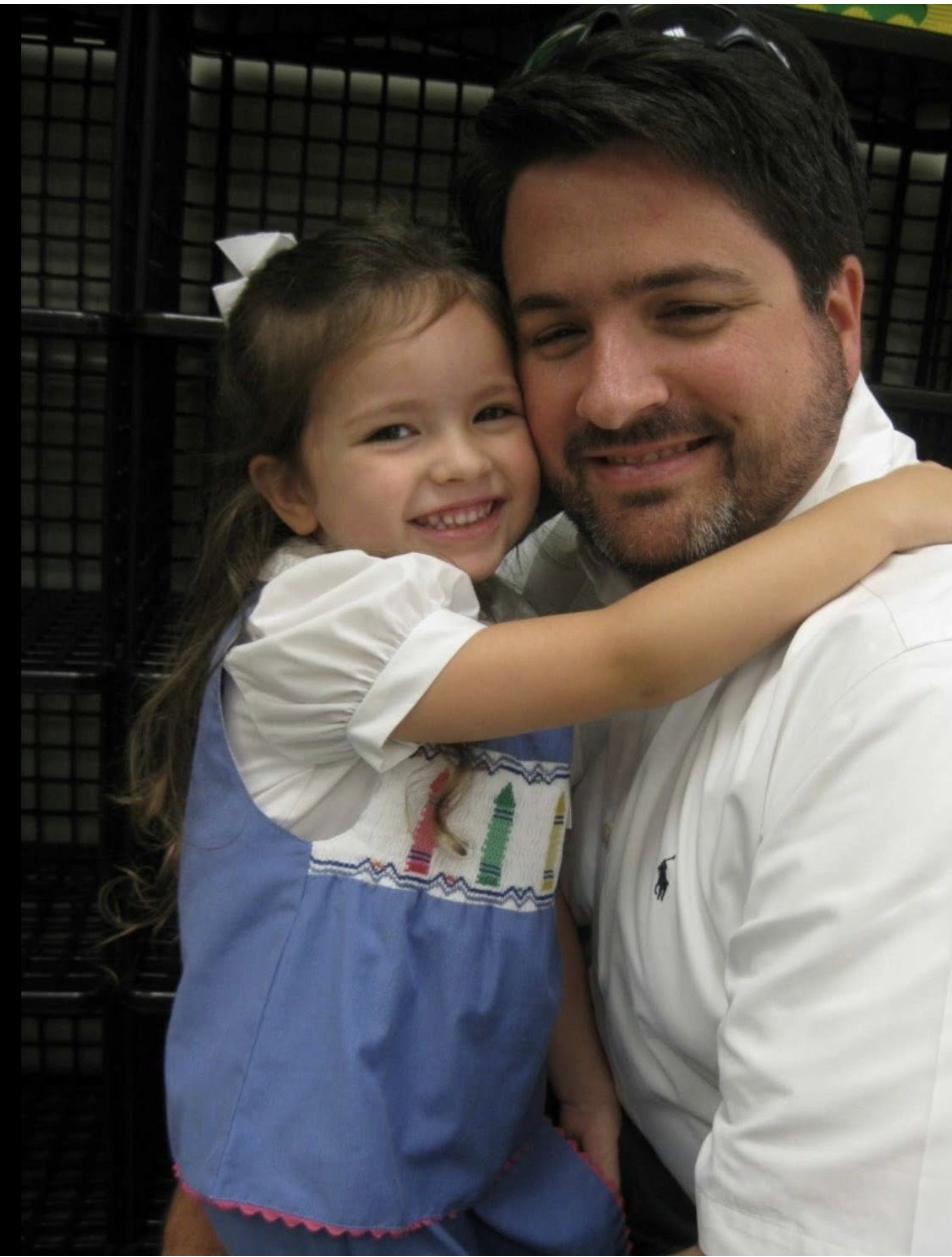 A young girl hugging a man, both smiling warmly, in an indoor setting with black shelving in the background.