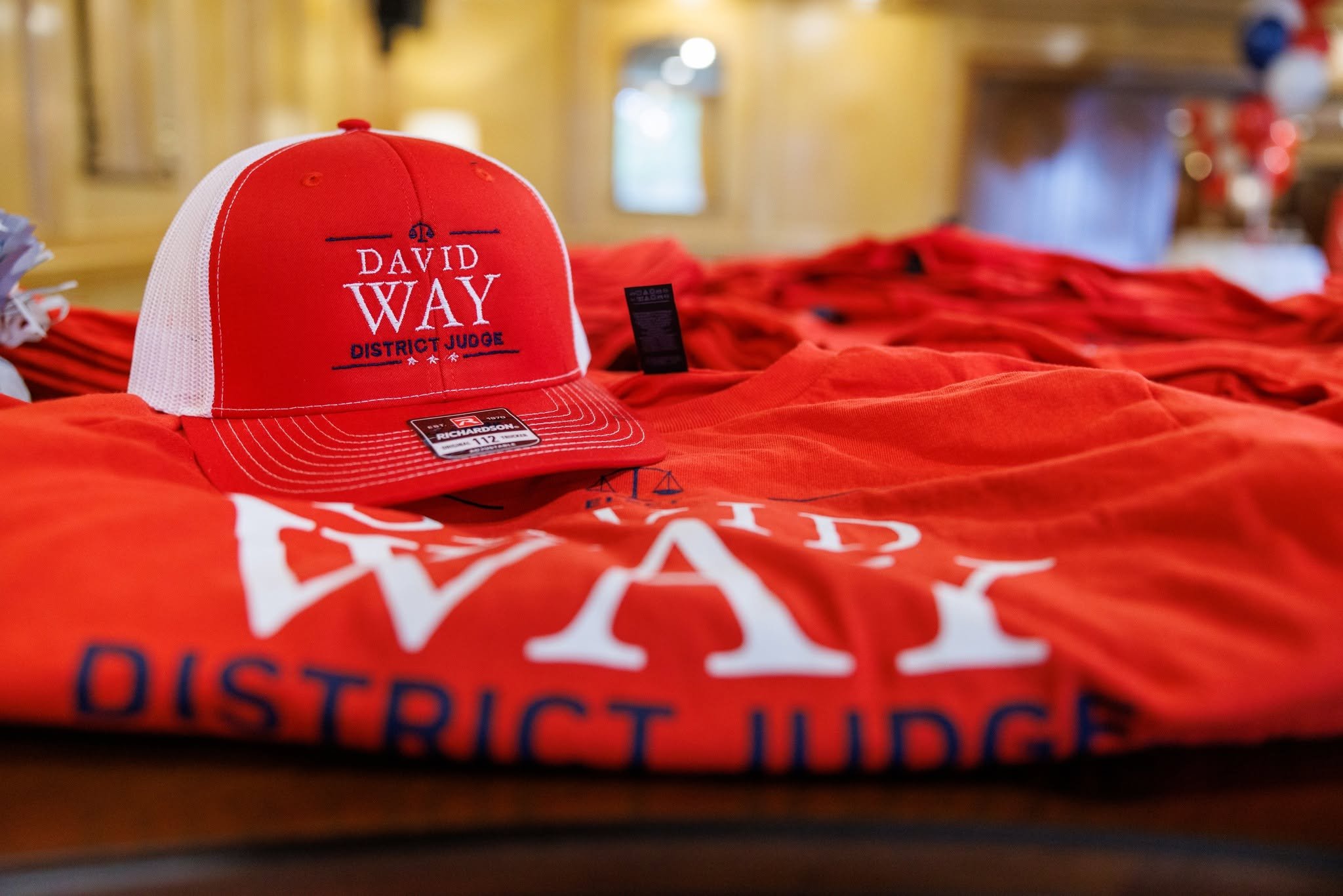 A red and white trucker hat with embroidered text reading "David Way District Judge" placed on top of red shirts with partial text visible. The background appears to be a blurred indoor setting.