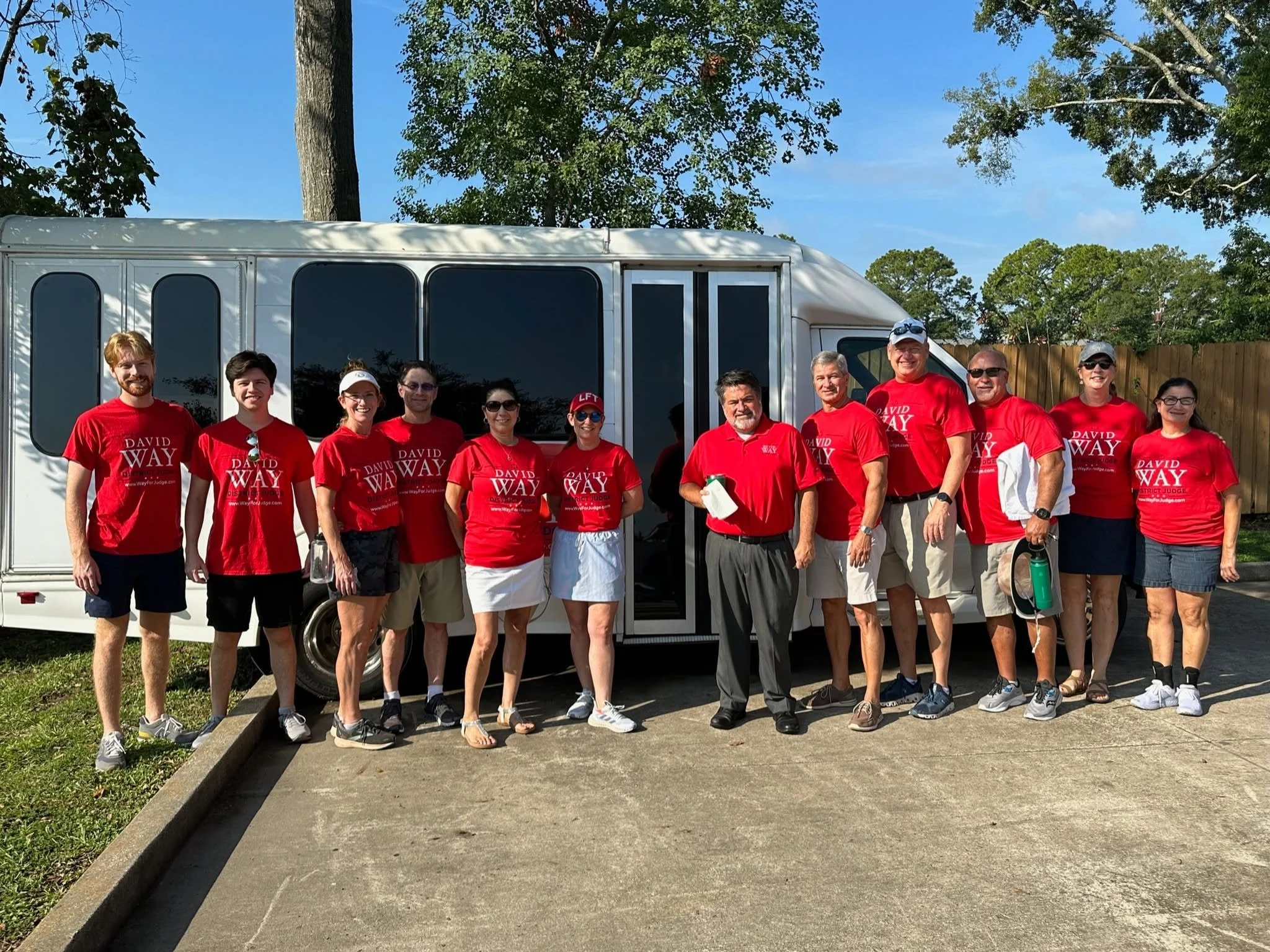 A group of people standing in front of a white shuttle bus, wearing red t-shirts with white text, outdoors on a sunny day with a blue sky and trees in the background.