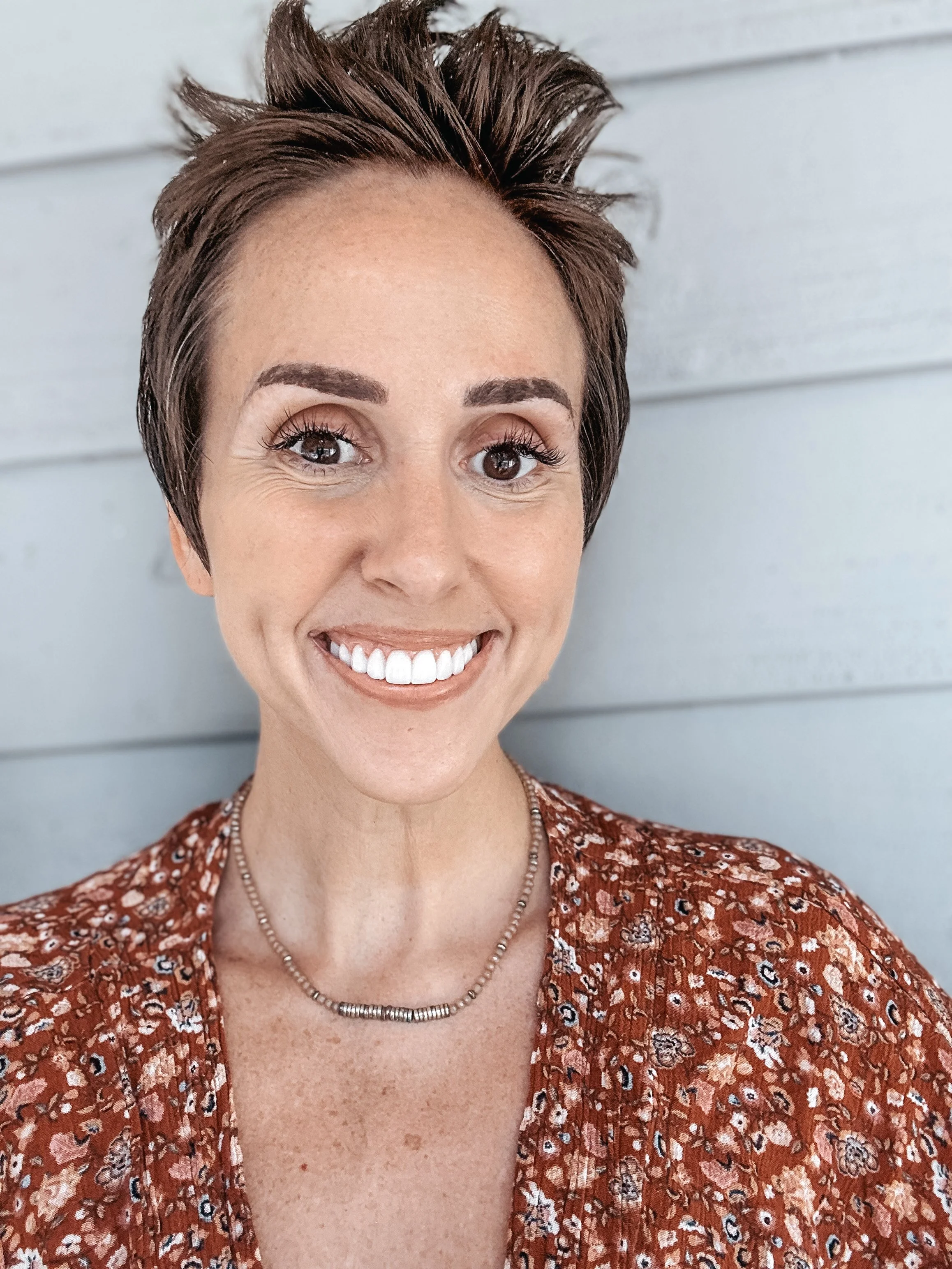A smiling woman with short, styled brown hair, wearing a patterned reddish-brown blouse and a beaded necklace, standing in front of a light blue wooden wall.