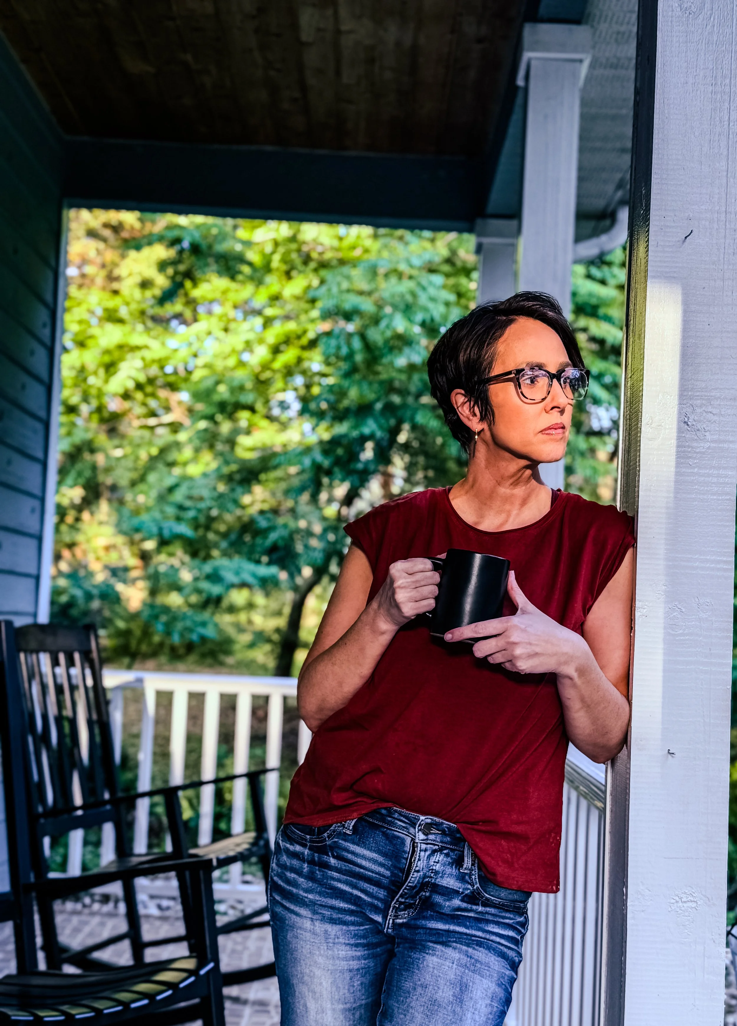 A woman with short dark hair and glasses standing on a porch, holding a black mug, looking to her right with a contemplative expression, in front of a background of green trees.