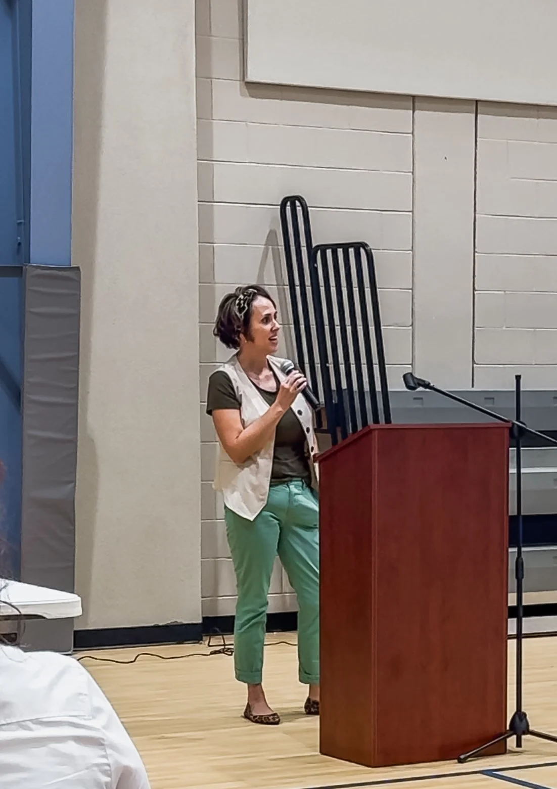 A woman with short dark hair, wearing a leopard print headband, a beige sleeveless vest over a brown shirt, teal pants, and leopard print shoes, stands next to a wooden podium and speaks into a microphone.