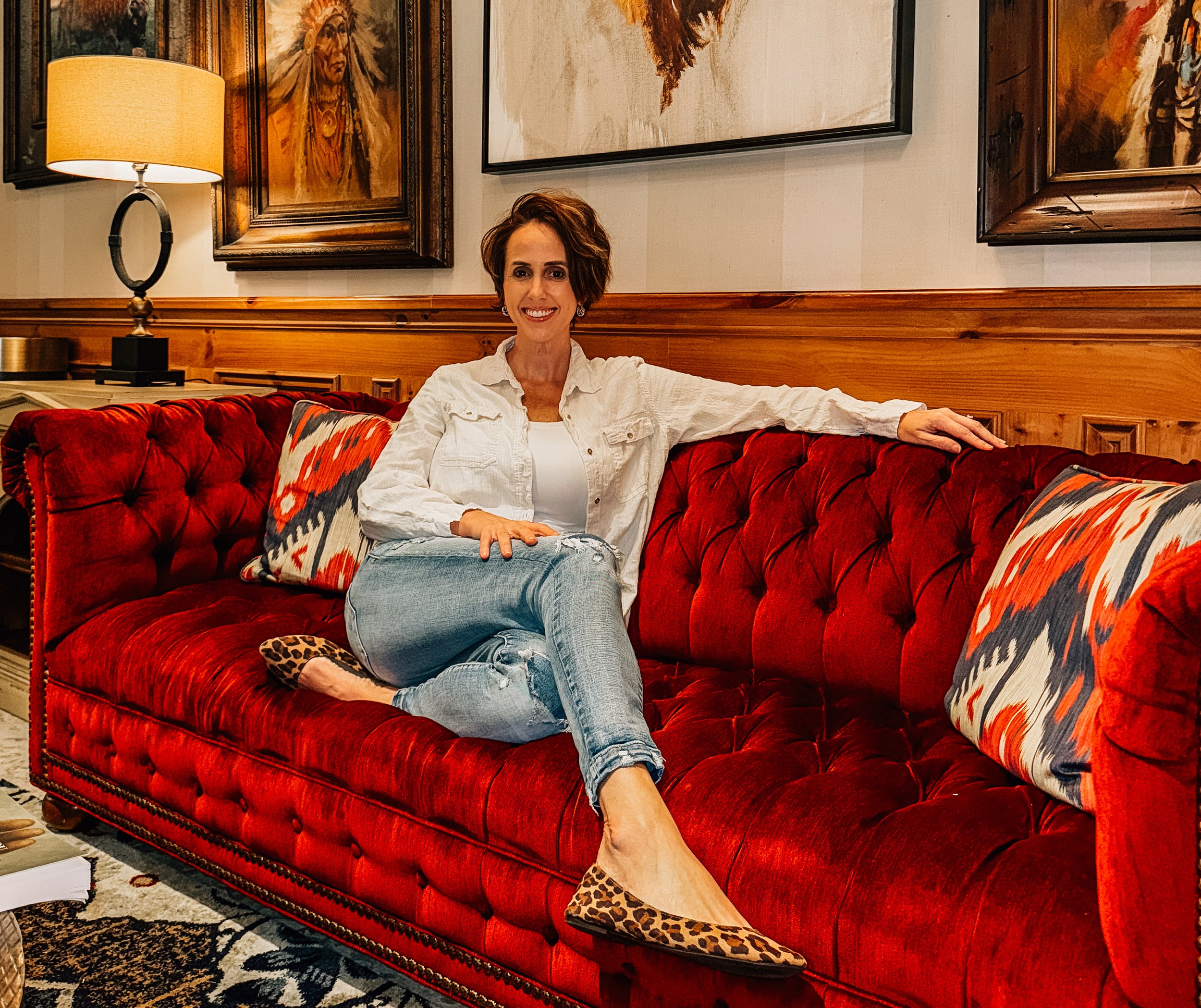 A woman with short brown hair smileing and sitting on a red velvet couch in a living room, wearing a white shirt, light blue jeans, and leopard print flats. There are colorful pillows and framed artwork on the wall behind her.