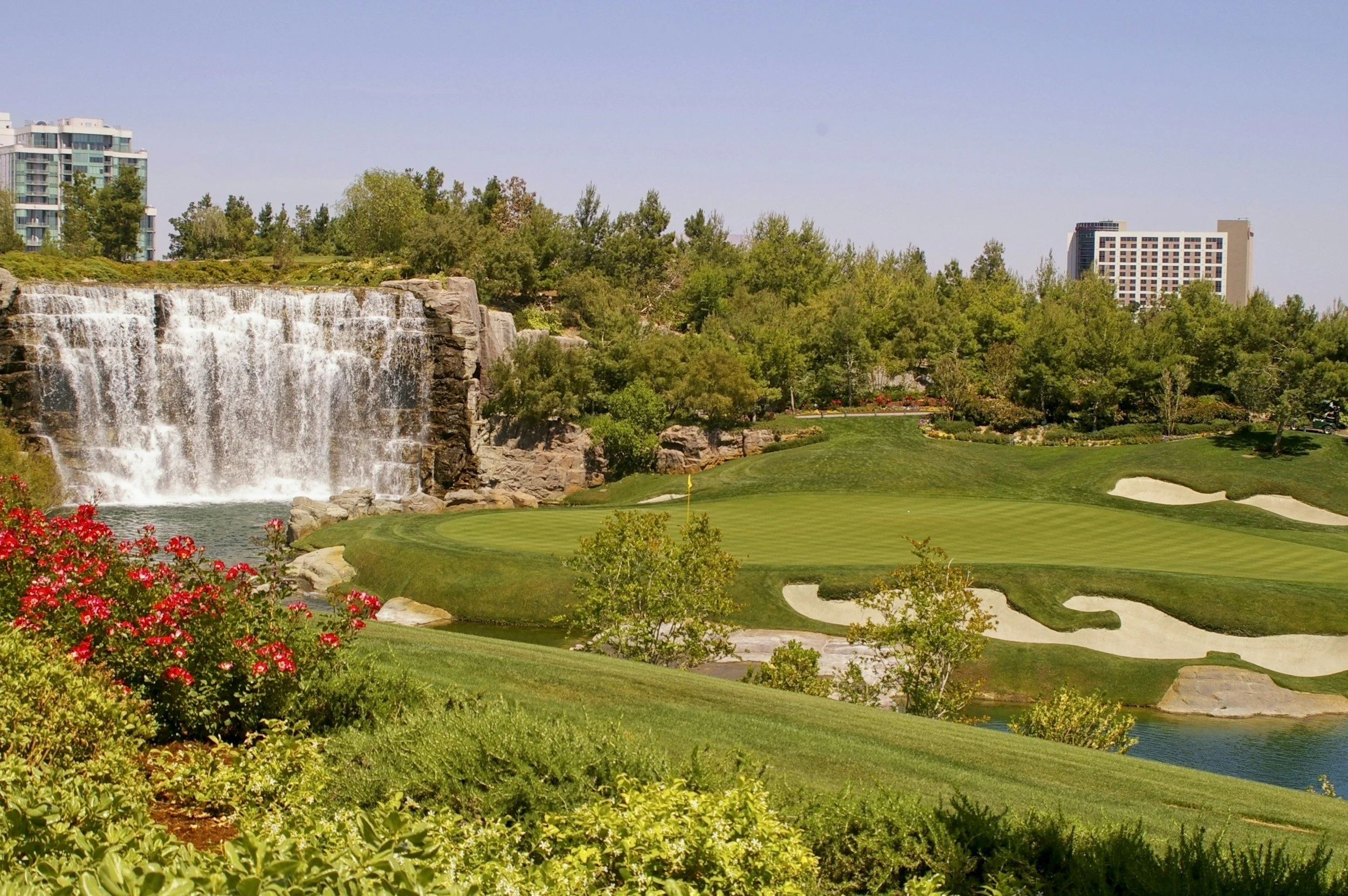 A scenic golf course with a waterfall on the left, surrounded by lush green trees and colorful flowers. Tall buildings are visible in the background under a clear blue sky.