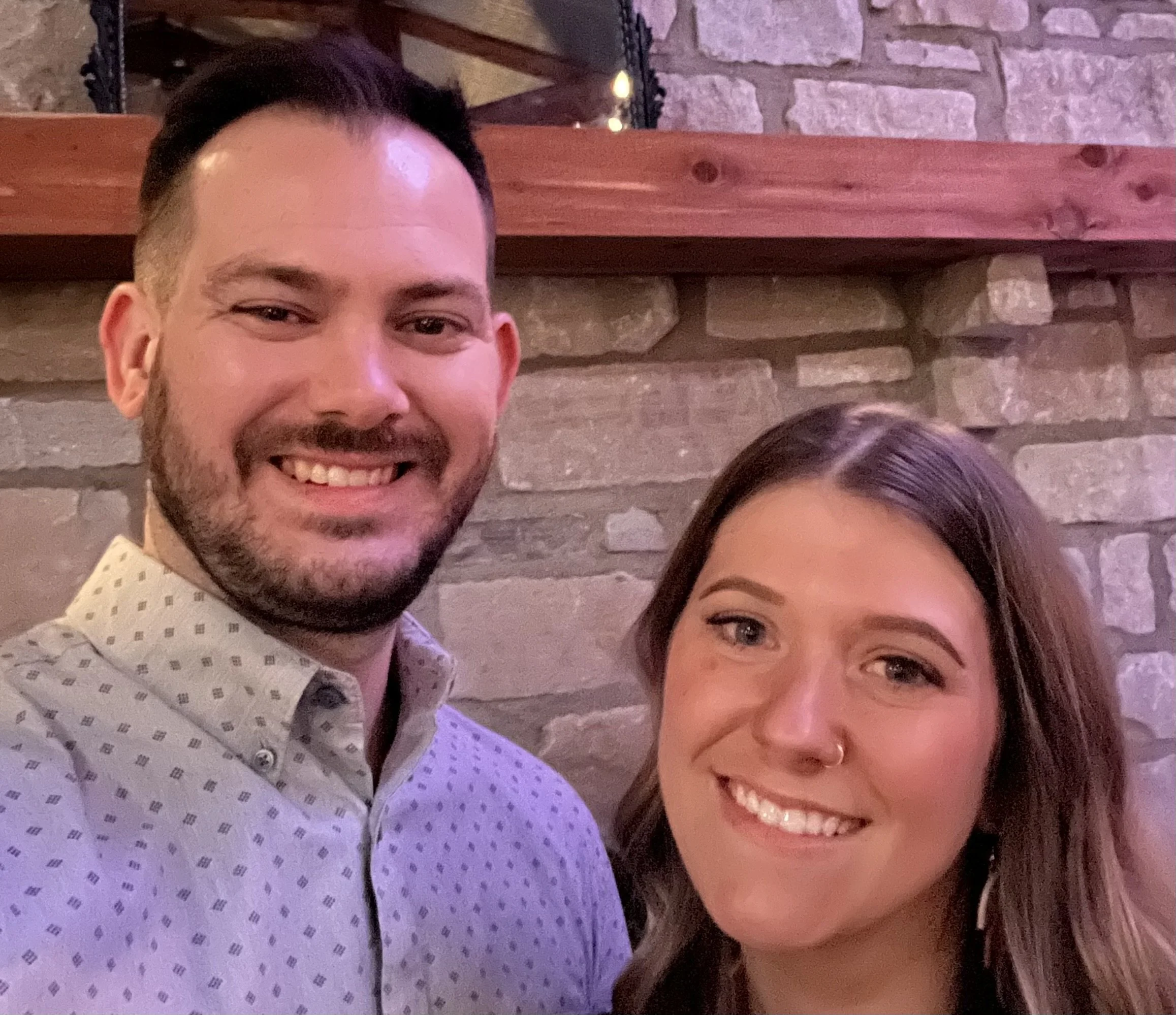 A smiling man and woman are taking a selfie together in a setting with a brick wall and wooden shelf behind them.