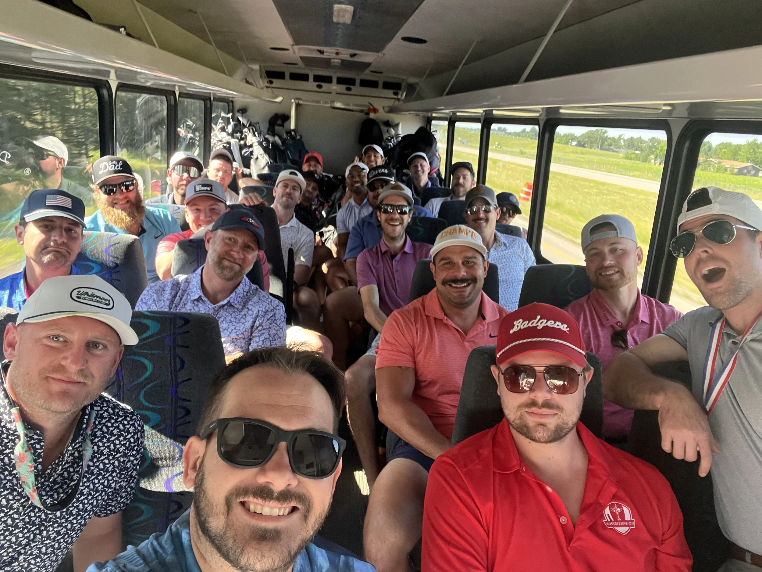 A group of men on a bus, dressed in casual golf attire, smiling and taking a selfie, with golf bags and equipment in the background.