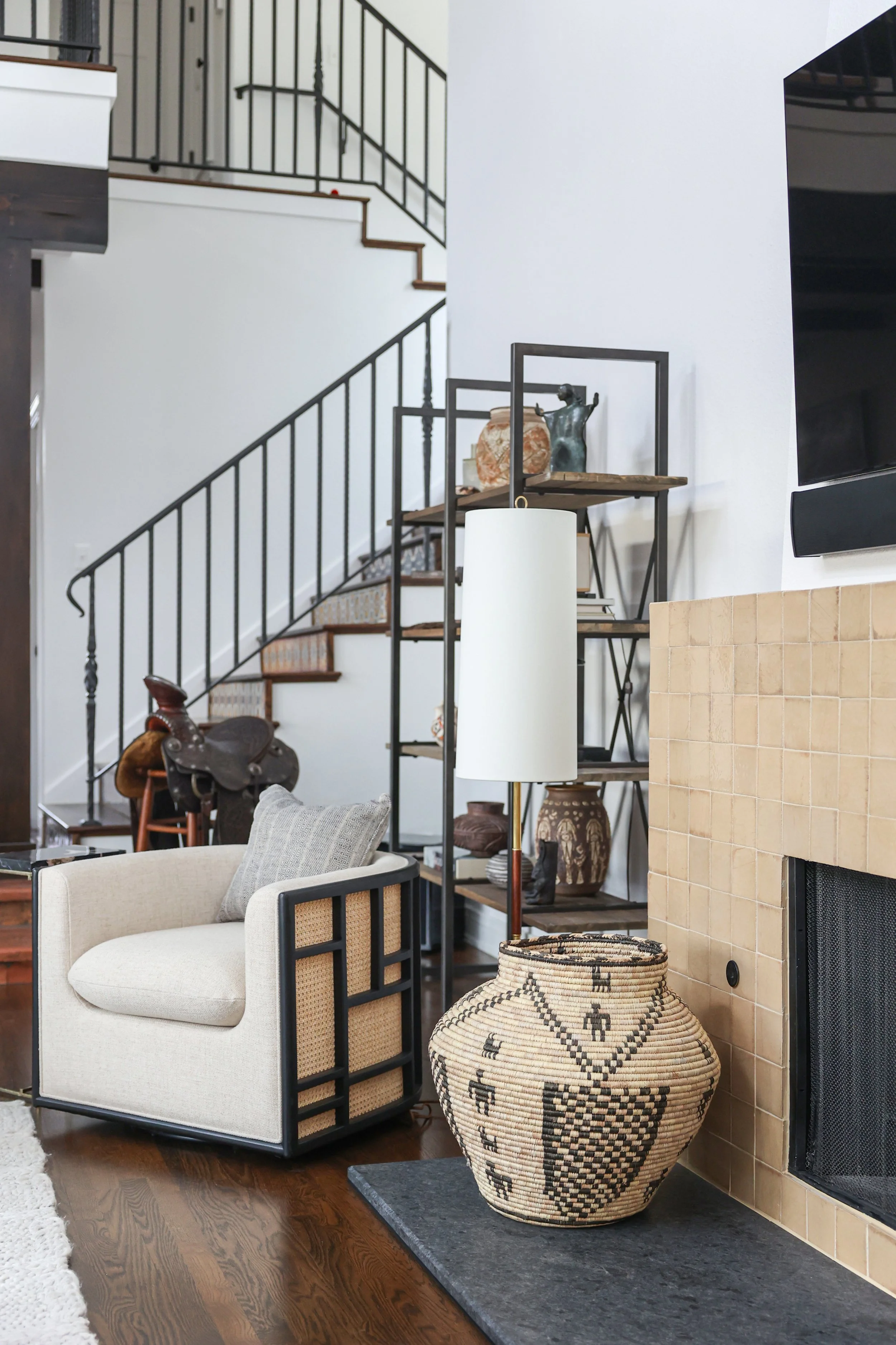 Living room in Modern Spanish Revival design style with art and collectibles, handmade tile on the fireplace, and a black metal shelving unit with decorative items, with a staircase in the background.
