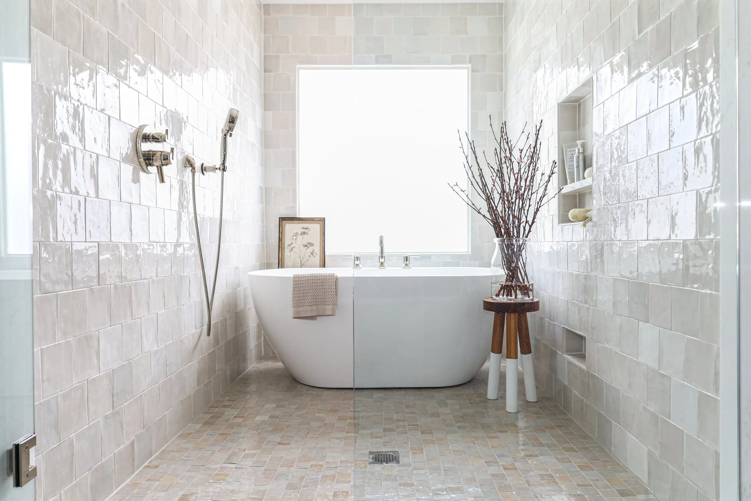 A modern bathroom with a white bathtub, large window, and beige tiled walls and floor. There are toiletries on a built-in shelf and a large vase with bare branches on a wooden stand. A framed picture leans against the bathtub, and a towel hangs over the tub's edge.