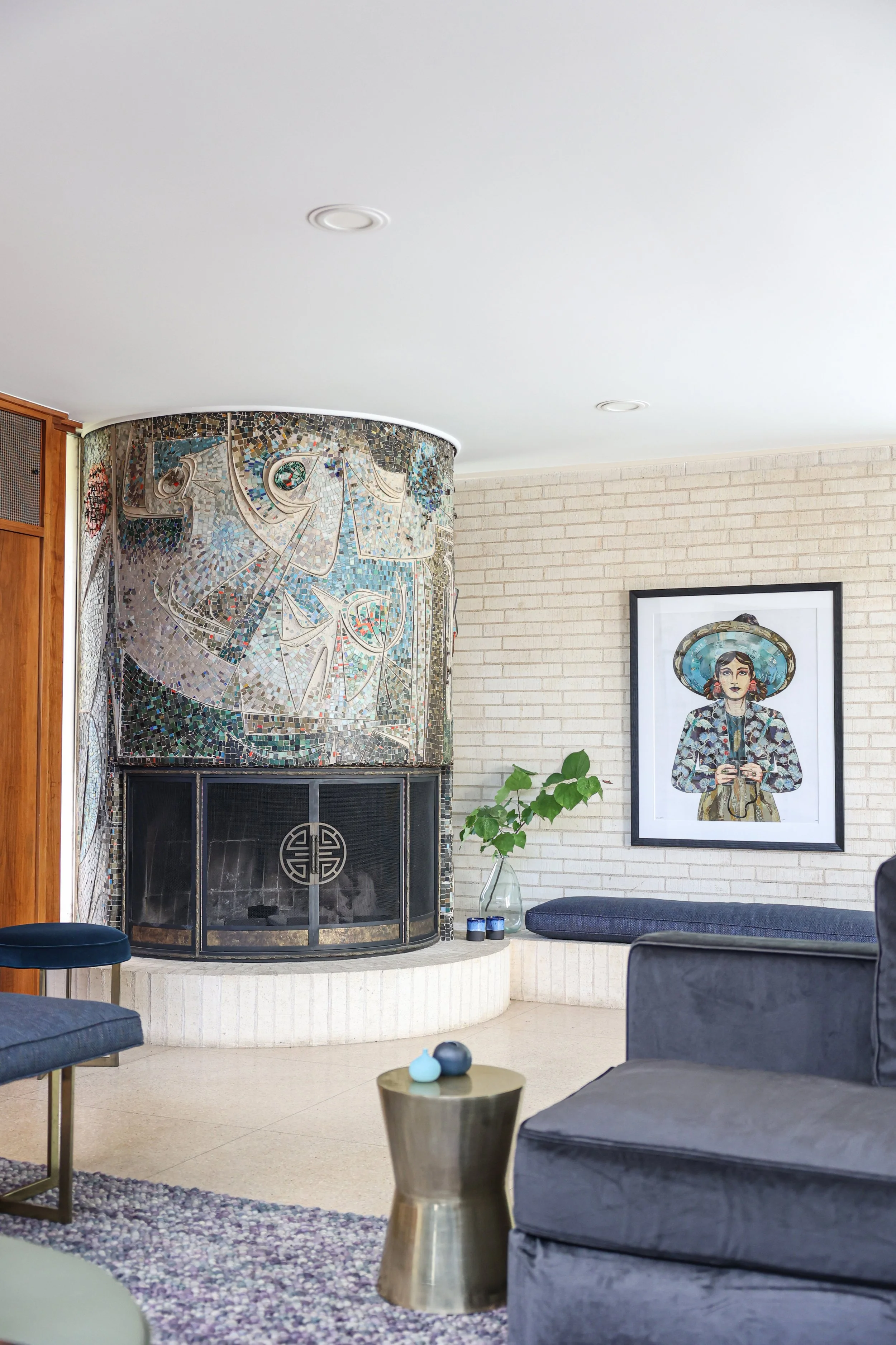 Living room with a white brick wall, a framed colorful illustration of a woman, a glass vase with green leaves, a gray sofa, and a side table with a gold base and small decorative objects. There is a tiled fireplace with an artistic design.
