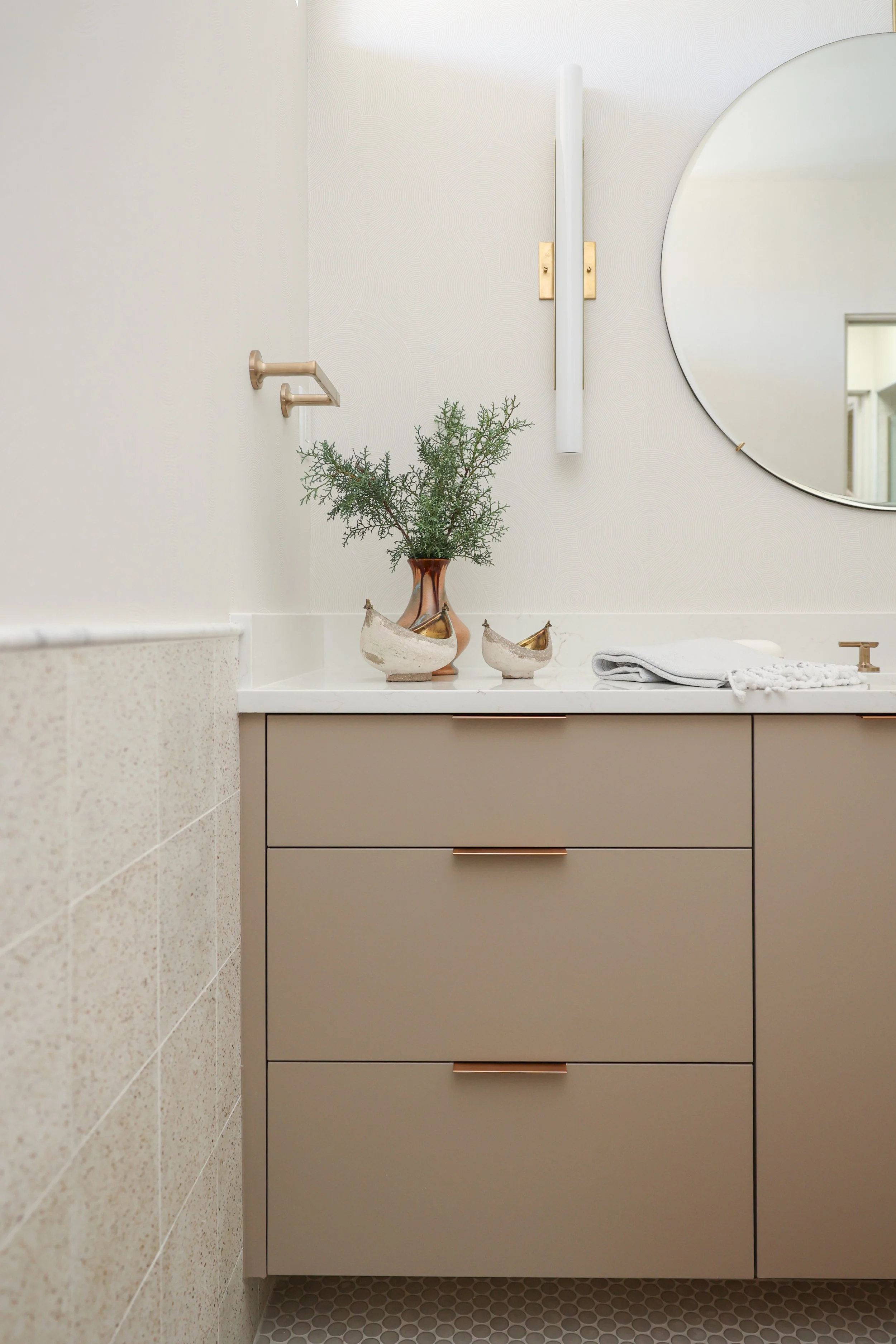Bathroom vanity with a beige cabinet, two drawers with rose gold handles, a white countertop with decorative vases, a folded white towel, and a round mirror on a white textured wall.