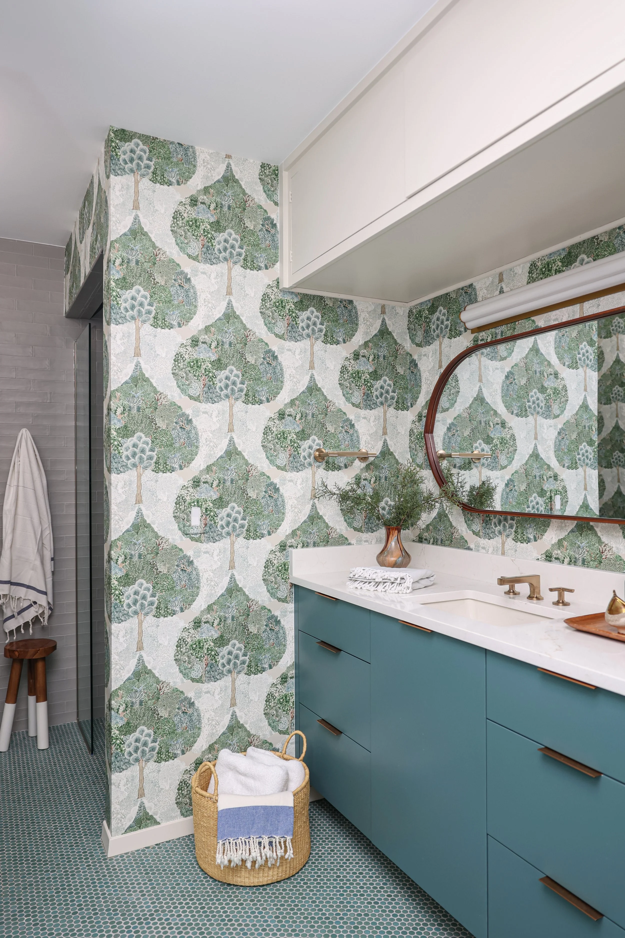A bathroom with a green and white patterned wallpaper, a white granite countertop with a blue cabinet, a large round mirror, a wicker basket with towels, and a towel hanging on a hook. The floor has small hexagonal tiles.