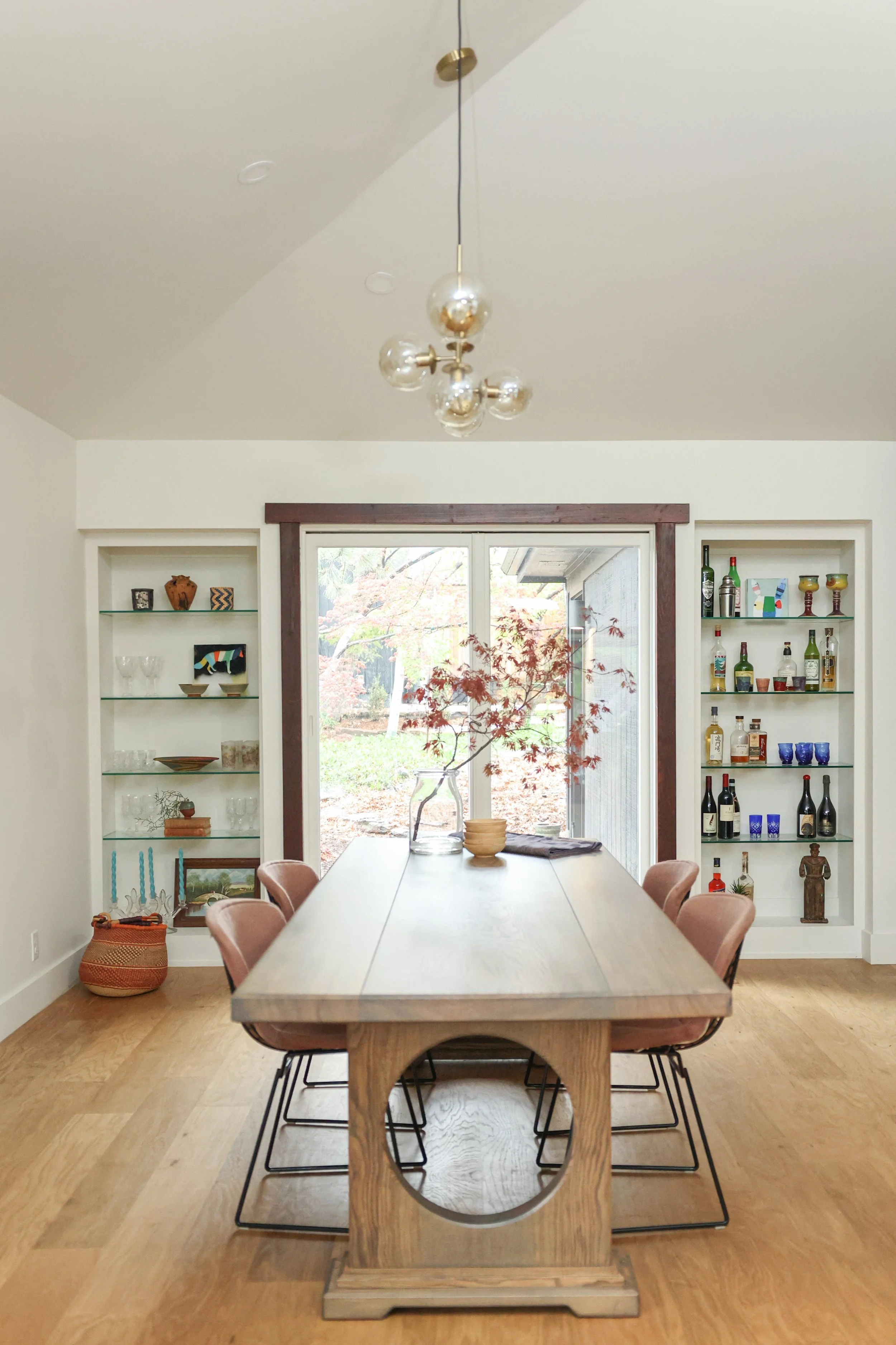 A dining room with a wooden table and six pink upholstered chairs. Behind the table are built-in shelves with decorative items and glasses. A window shows a garden outside. A modern pendant light hangs above the table.