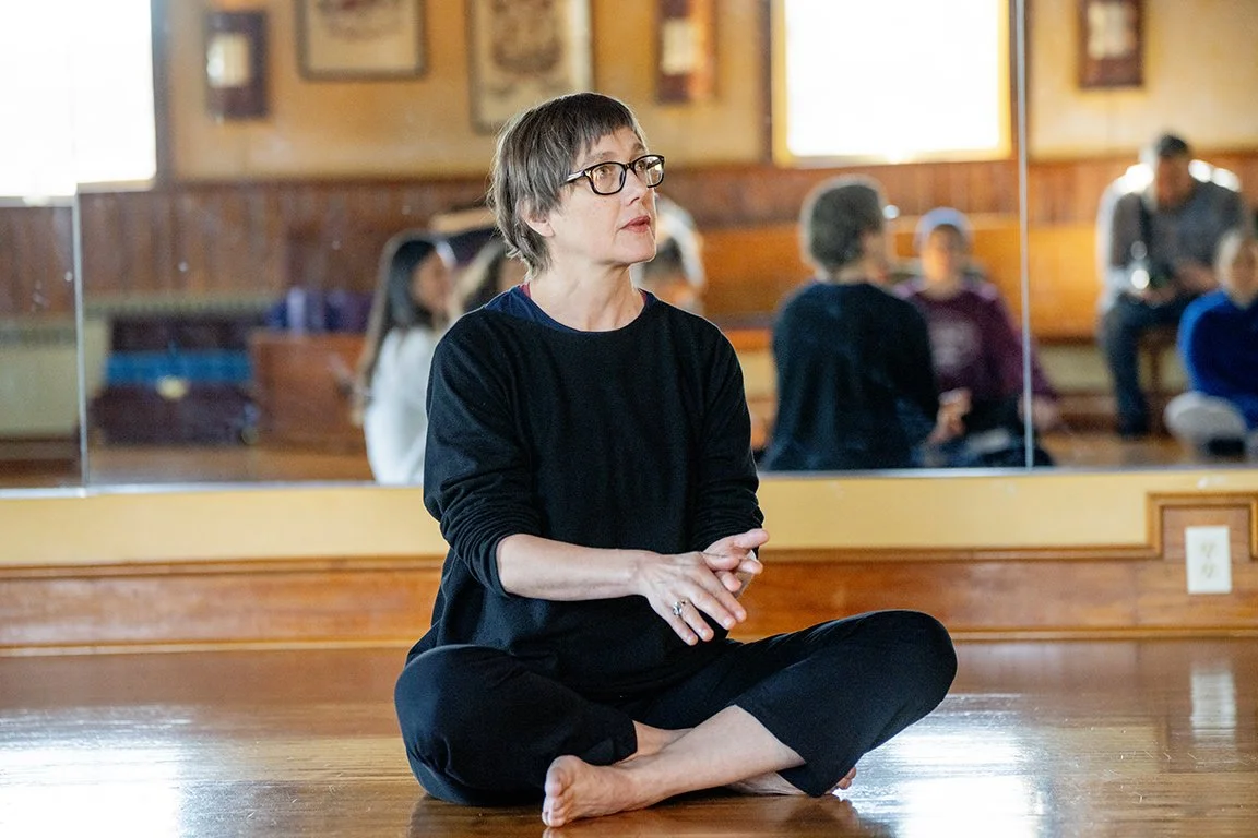 Liesje Smith is wearing black and has short blond hair and glasses. She is sitting on the floor and gesturing with her hands, indicating that she is teaching about chronic pain and Rolfing
