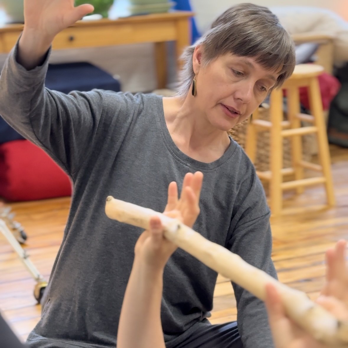 A woman with short hair, wearing a gray long-sleeve shirt, is engaging a Rolf Movement student, who is holding a wooden stick and moving it in an arc. The woman appears attentive and focused on the student's movement pathway.