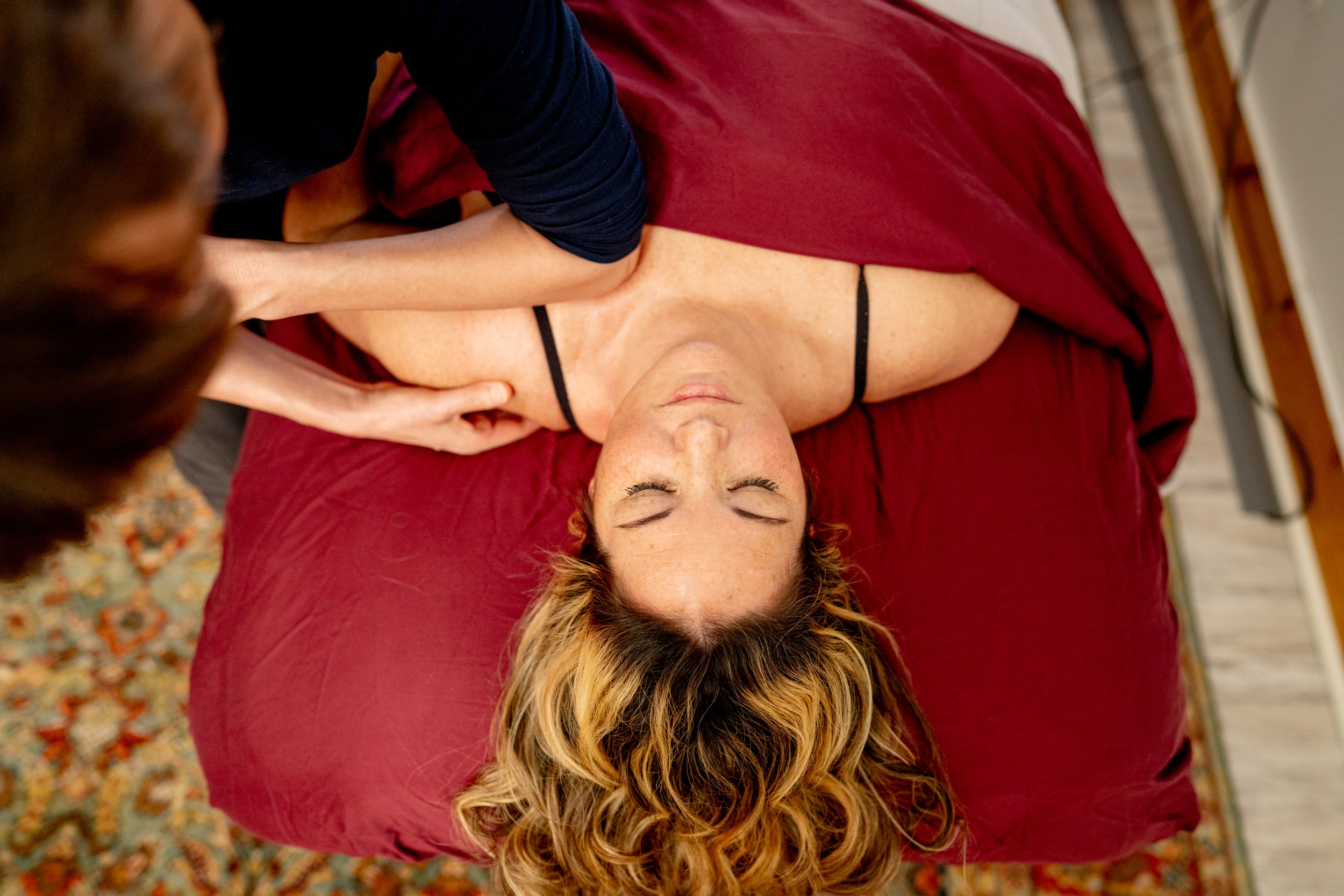 A woman lying on her back on a massage table, receiving Rolfing from a Roller. The woman has curly hair, is wearing a black top, and appears relaxed with closed eyes. The Rolfing is focused on her shoulder area.