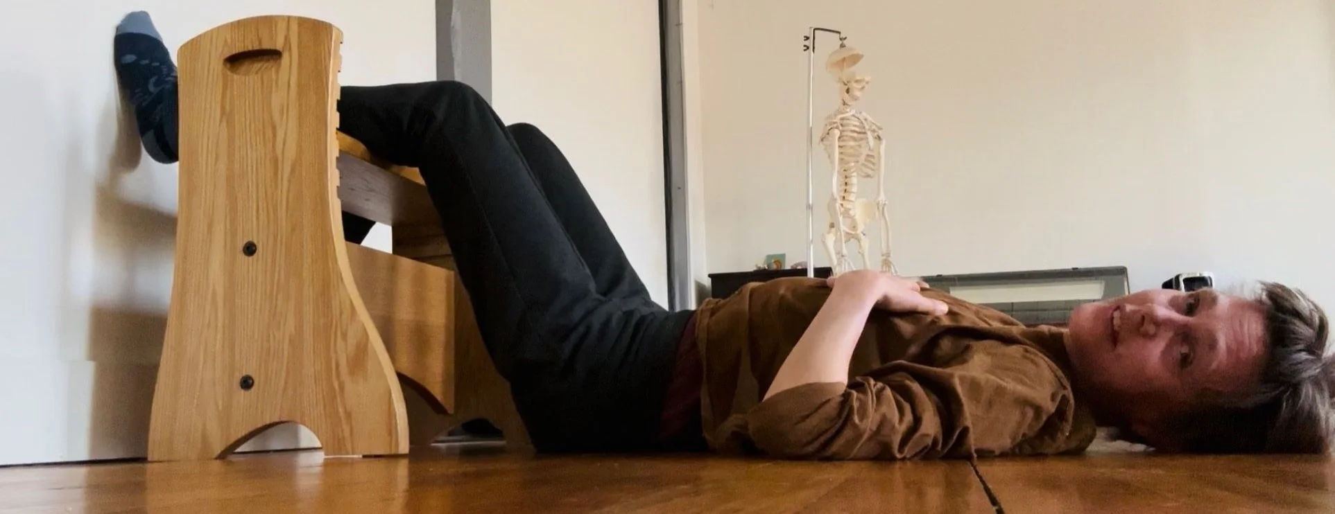 Liesje Smith  lying on their back on the floor with their legs elevated on a wooden bench, smiling at the camera. There is an anatomical skeleton model in the background.
