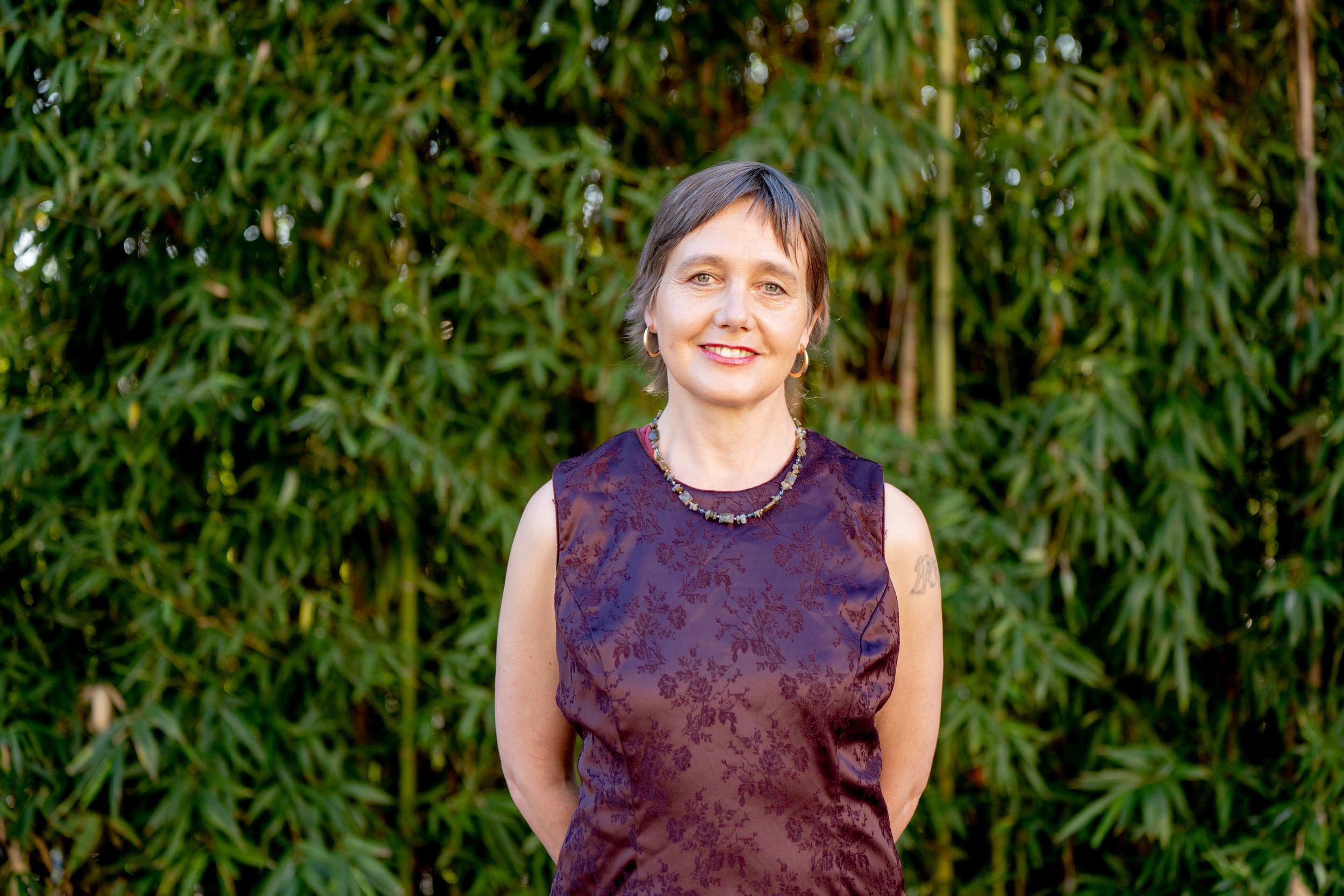 Liesje Smith with short brown hair smiling, wearing a sleeveless purple dress with a floral pattern, standing outdoors in front of green foliage.