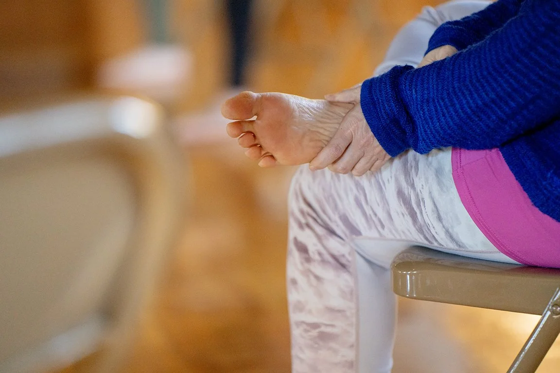 A person wearing a blue sweater and pink pants sitting on a beige chair, holding their stretched foot with their hand in a room with wooden flooring.