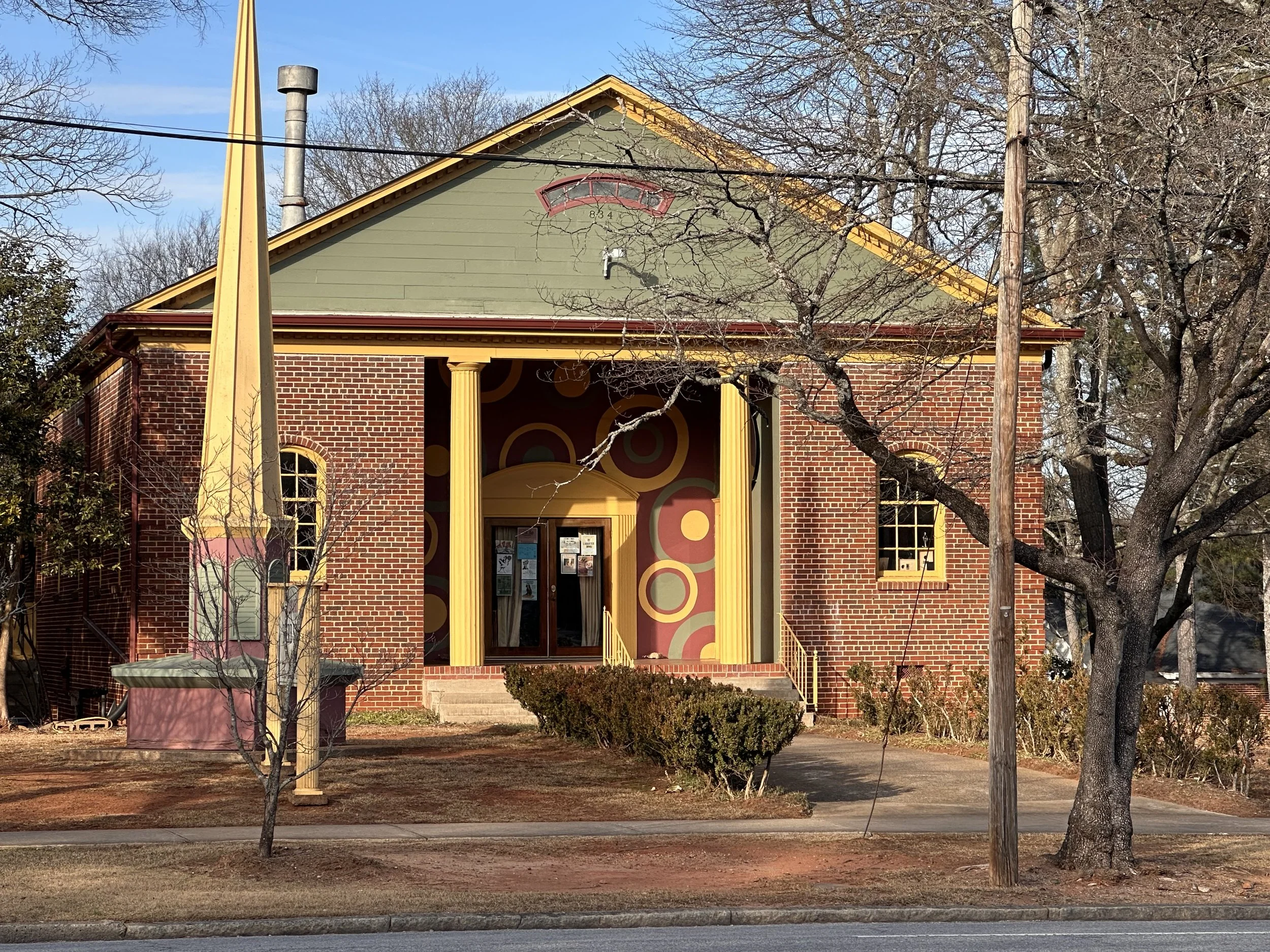 A colorful building with a brick facade, yellow columns, and a mural with circles behind the entrance. There are trees, bushes, and utility poles in the yard, with a sidewalk and street in the foreground.