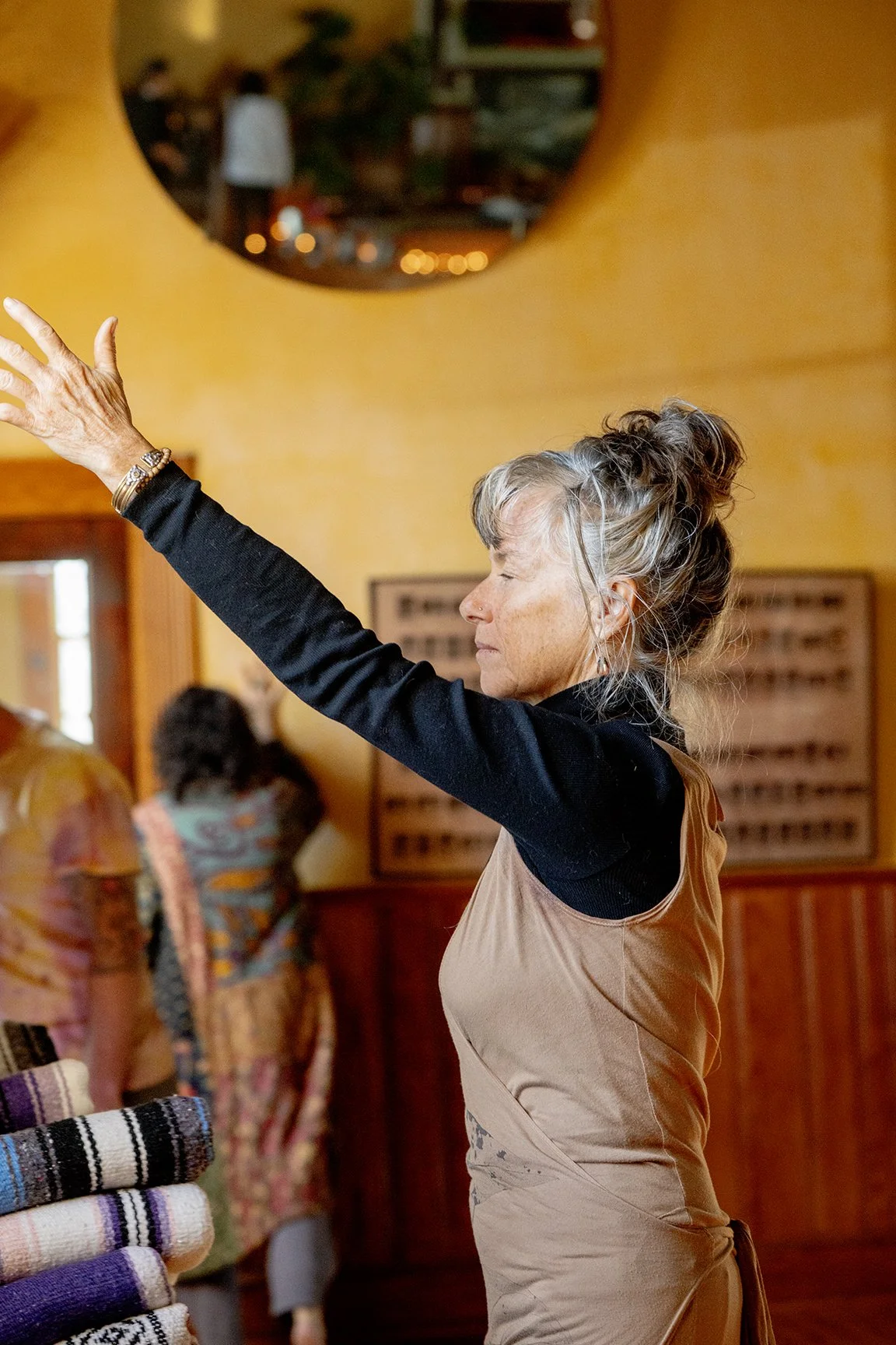 An elderly woman with gray hair tied up, wearing a black long-sleeve shirt and a beige apron, standing indoors with her eyes closed and arms raised, possibly practicing yoga or meditation.