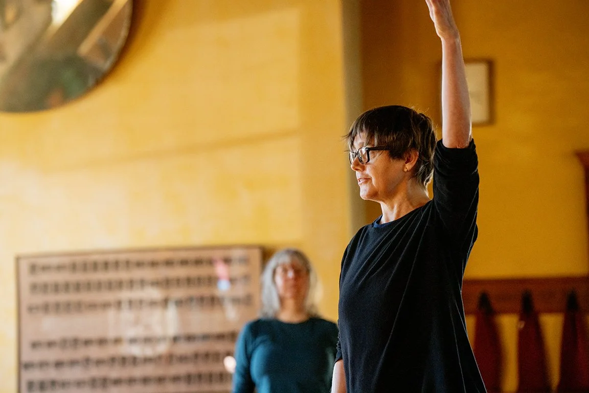 Liesje Smith wearing glasses and a black shirt is raising her right arm in a Rolf Movement class. Another woman with gray hair and glasses, wearing a teal shirt, is slightly out of focus in the background.