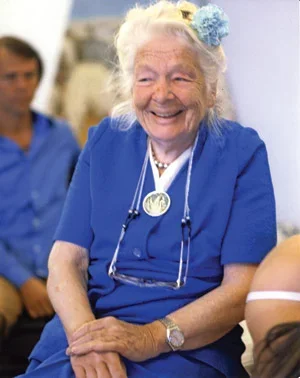 Dr. Ida P. Rolf, elderly woman wearing a blue dress with a carnation in her hair. She is smiling, perhaps laughing