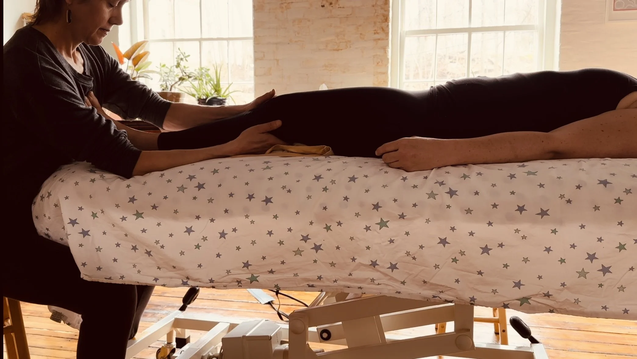 A person lying on a bed with a star-patterned bedsheet, receiving SourcePoint Therapy from Liesje Smith who is sitting at the end of the table with her hands on the client's knee in a well-lit room with large windows and potted plants.