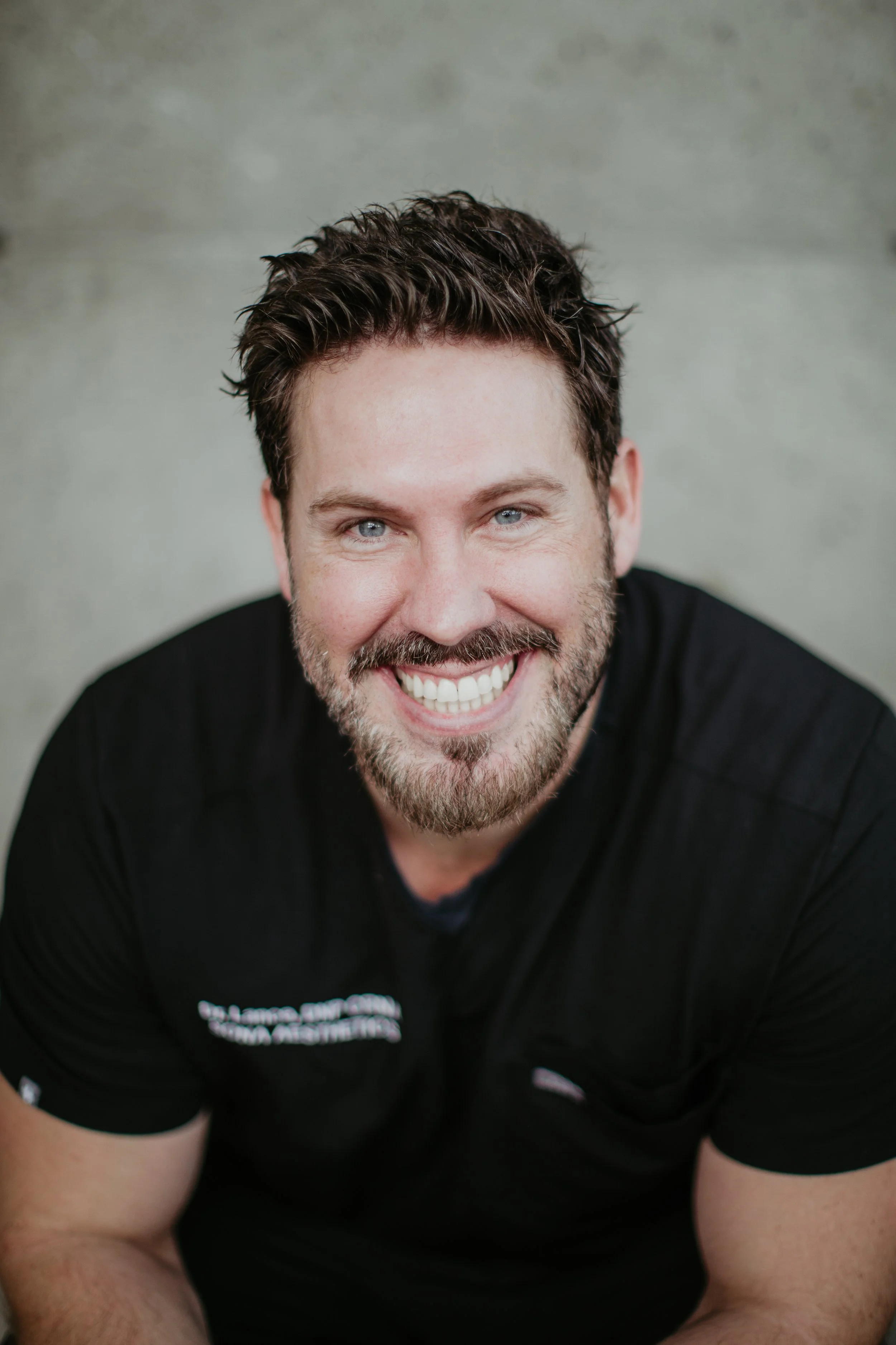 Lance Kennedy ARNP CRNA leaning forward and smiling in Sona Aesthetics scrubs at Sona Medical Aesthetics Newcastle WA