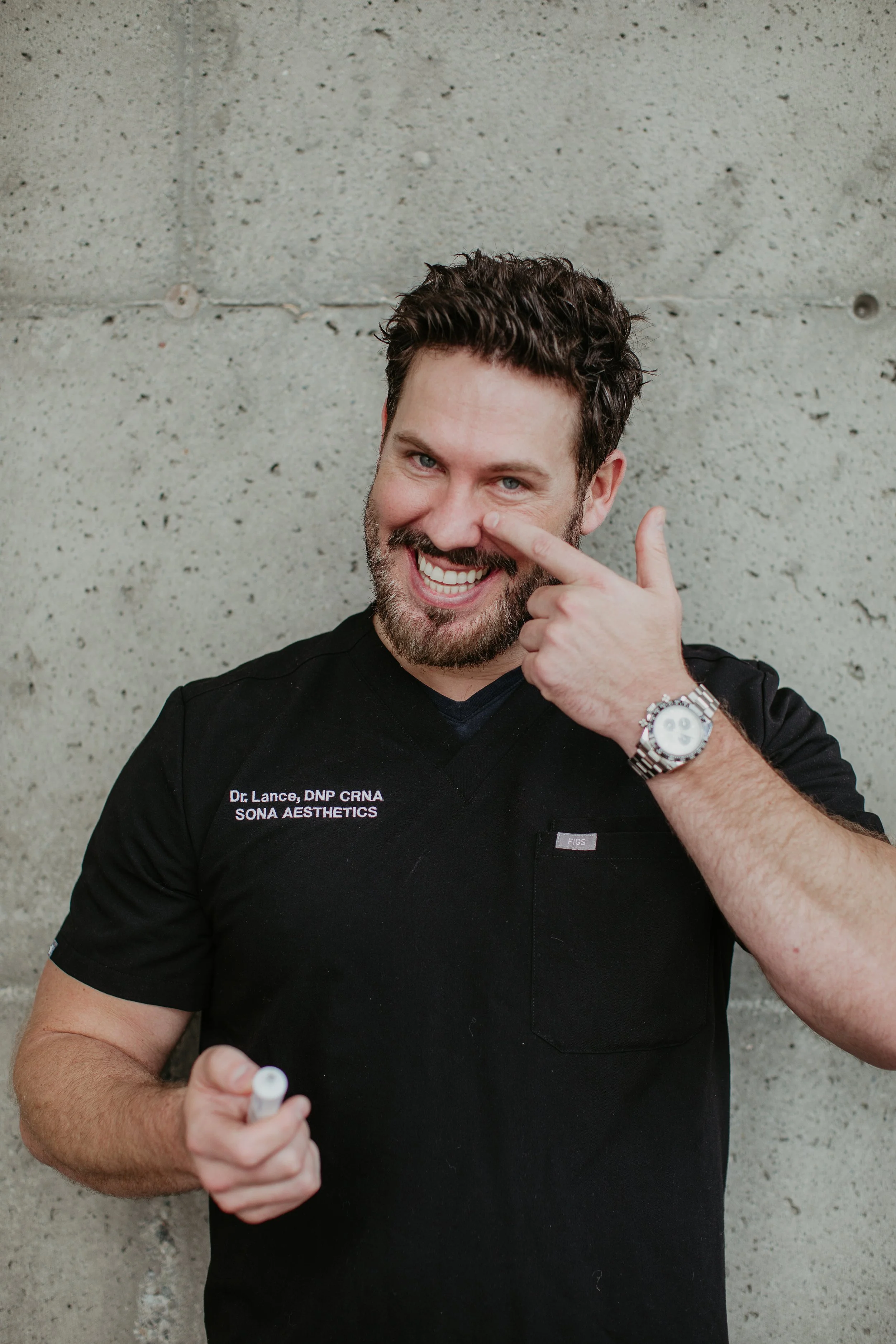 Lance Kennedy DNP CRNA pointing to under-eye area while holding an injectable vial at Sona Medical Aesthetics Newcastle WA