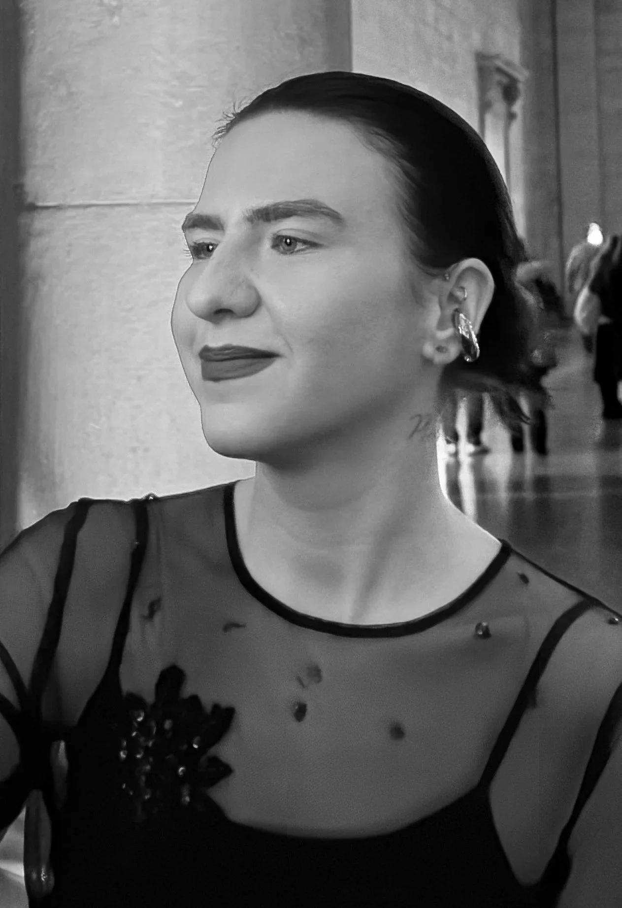 A woman with dark hair and dark lipstick wearing a sheer top with floral embroidery in a black and white photo.