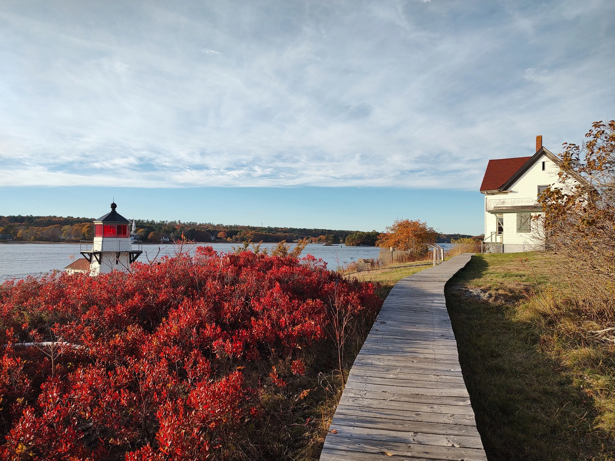 Small lighthouse behind red foliage and a board walk to a white building