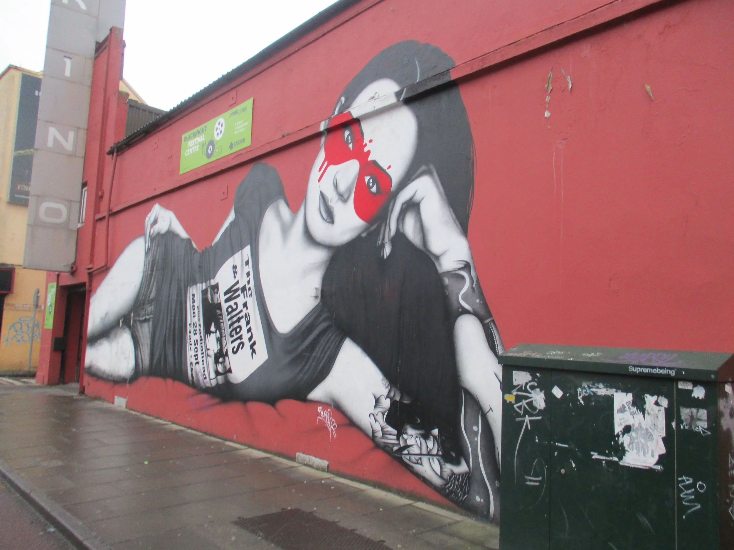 street art of a woman laying on her side in shorts and a t-shirt that says "The Frank & Walters." She is in black and white except for an abstract eye mask that surrounds her eyes in red. The background is red.