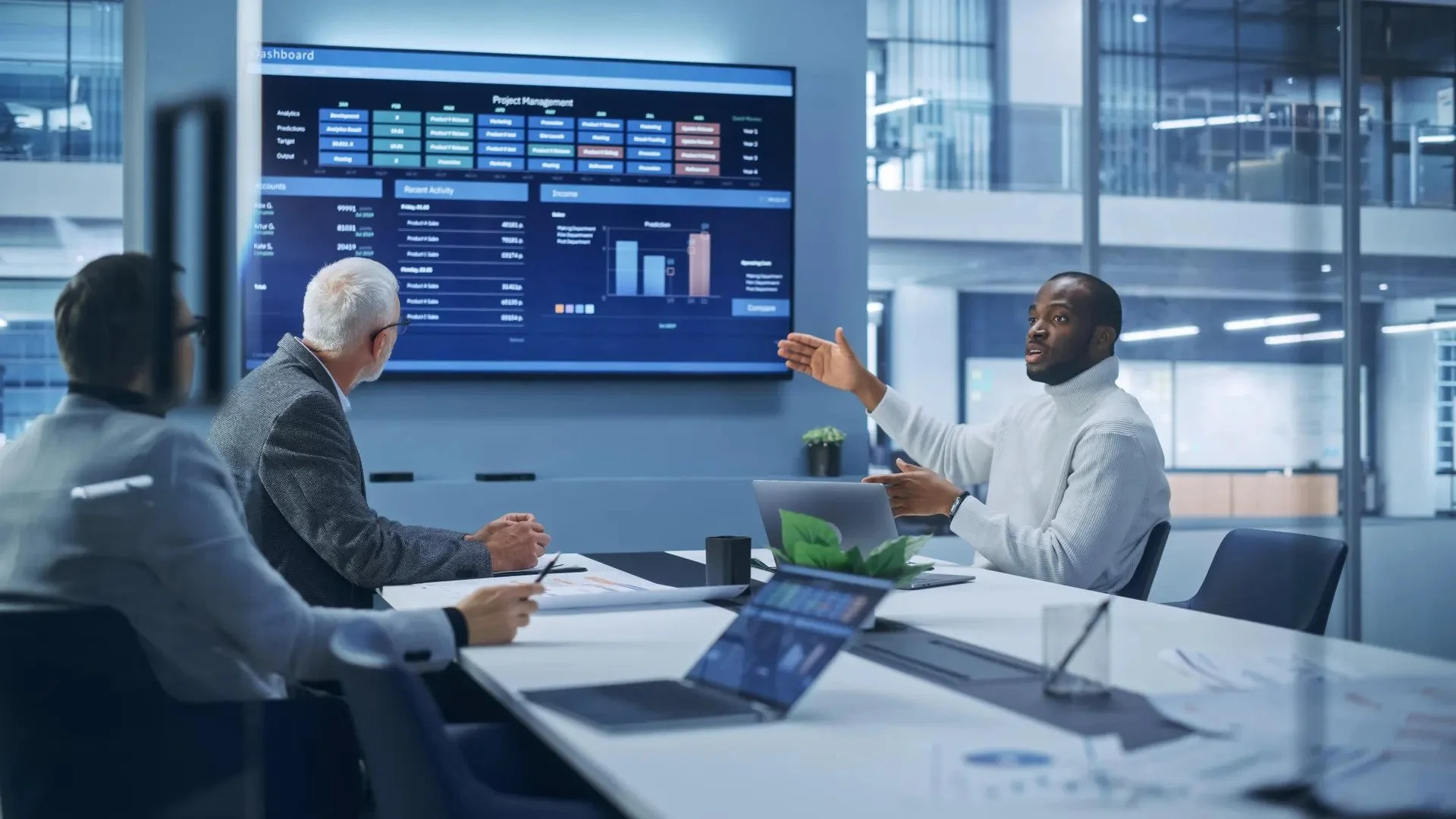 Business meeting in a modern conference room with three professionals discussing data and project management displayed on a large screen.